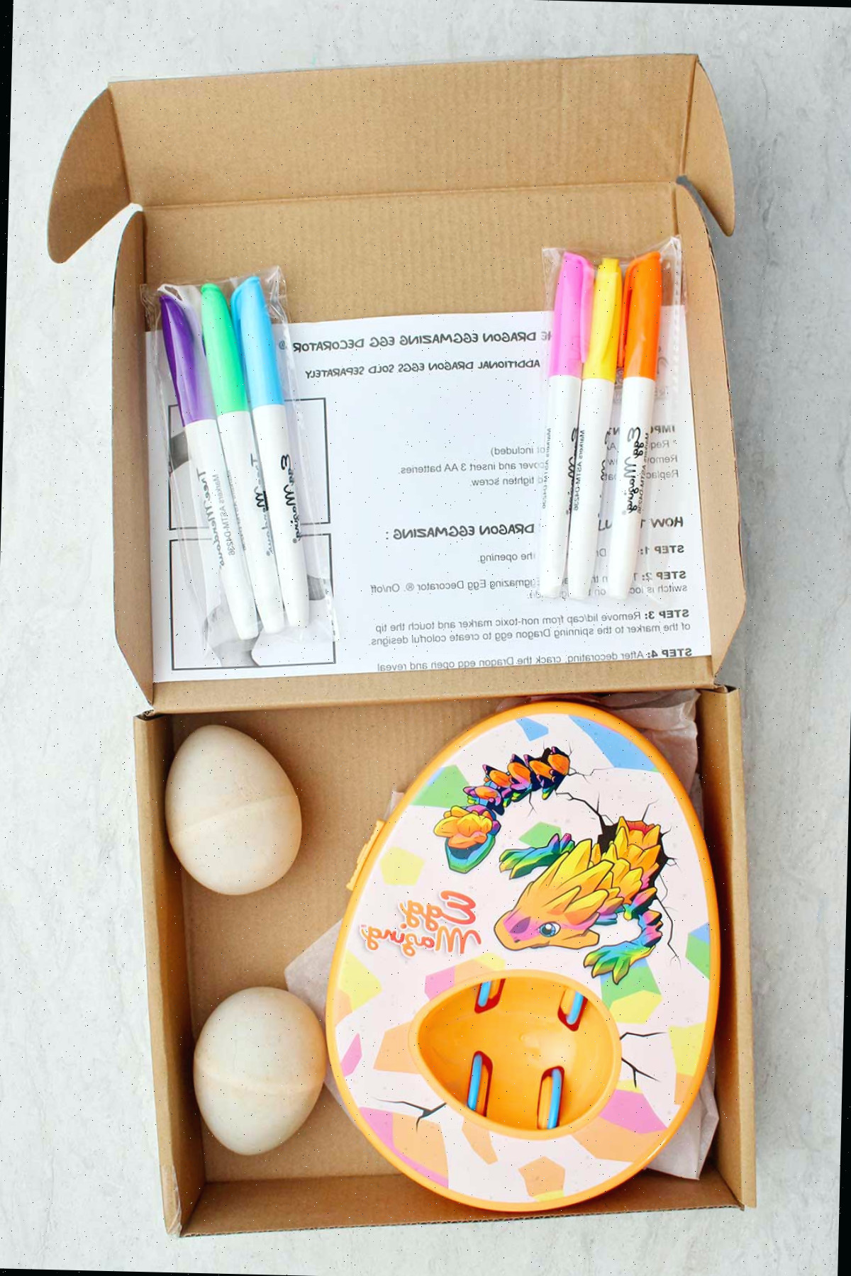Uma caixa cheia de um kit EggMazing, marcadores, instruções e ovos de madeira.