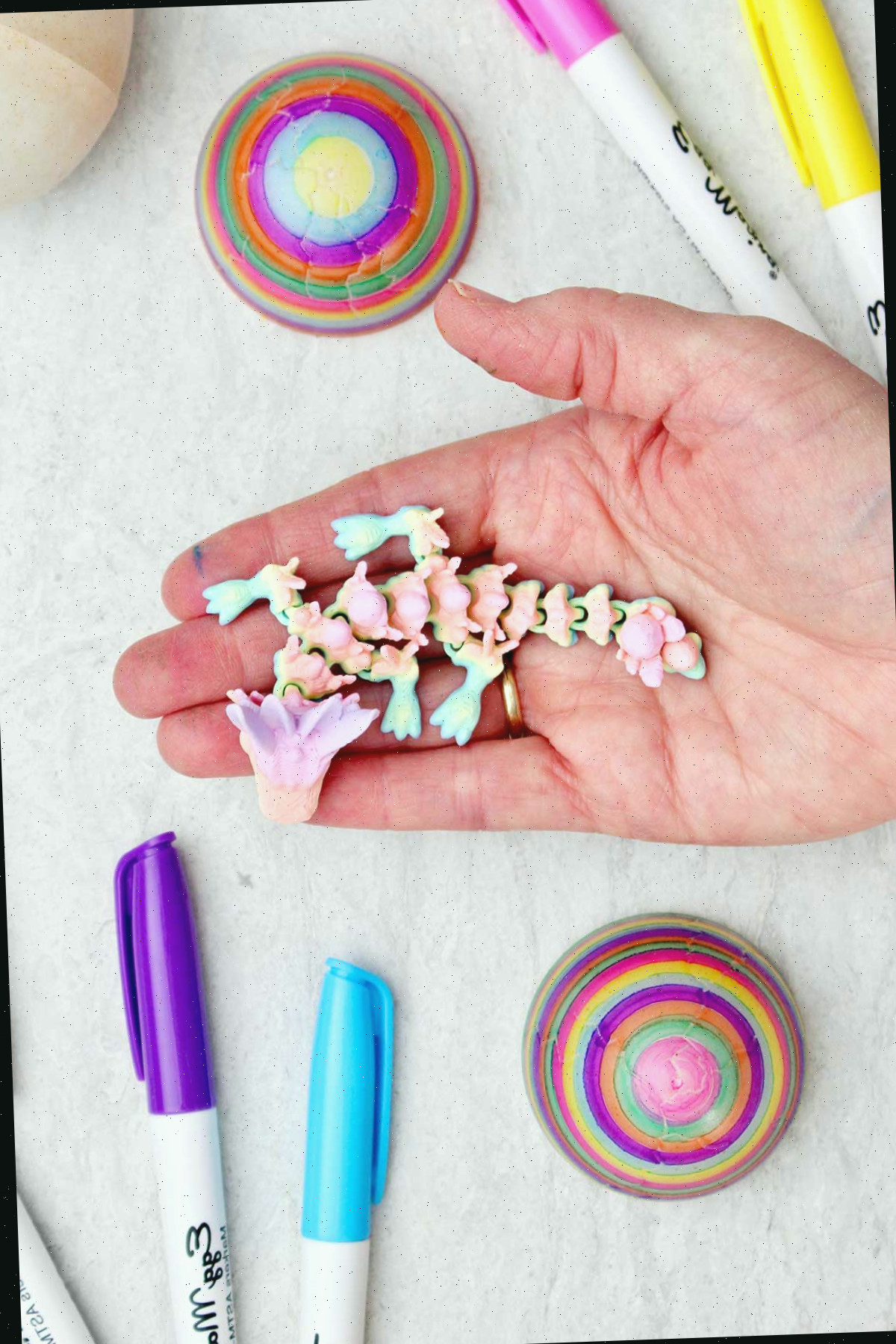 Uma pessoa segurando um dragão colorido impresso em 3D com dois ovos coloridos e canetas próximas.