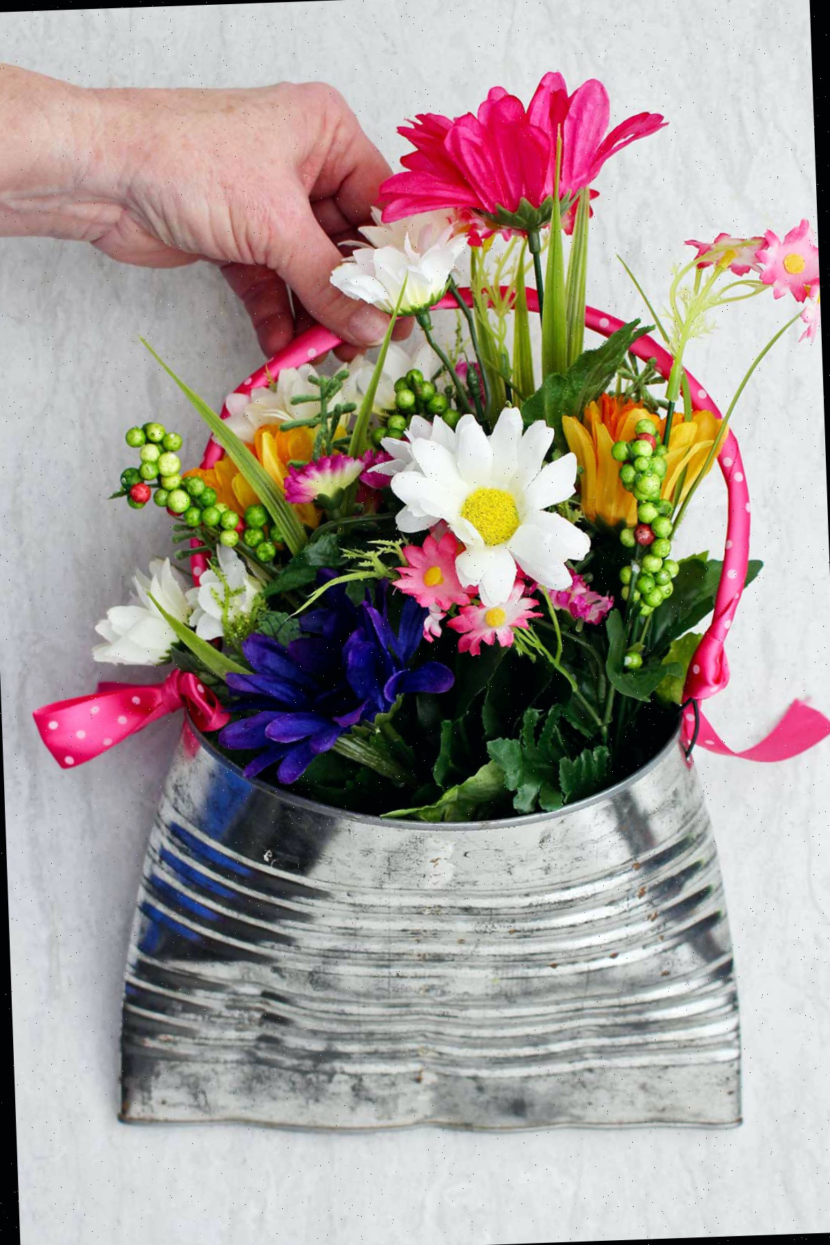 Uma pessoa segurando um vaso de lata reciclado completo, cheio de flores rosas, brancas e roxas com folhagens.