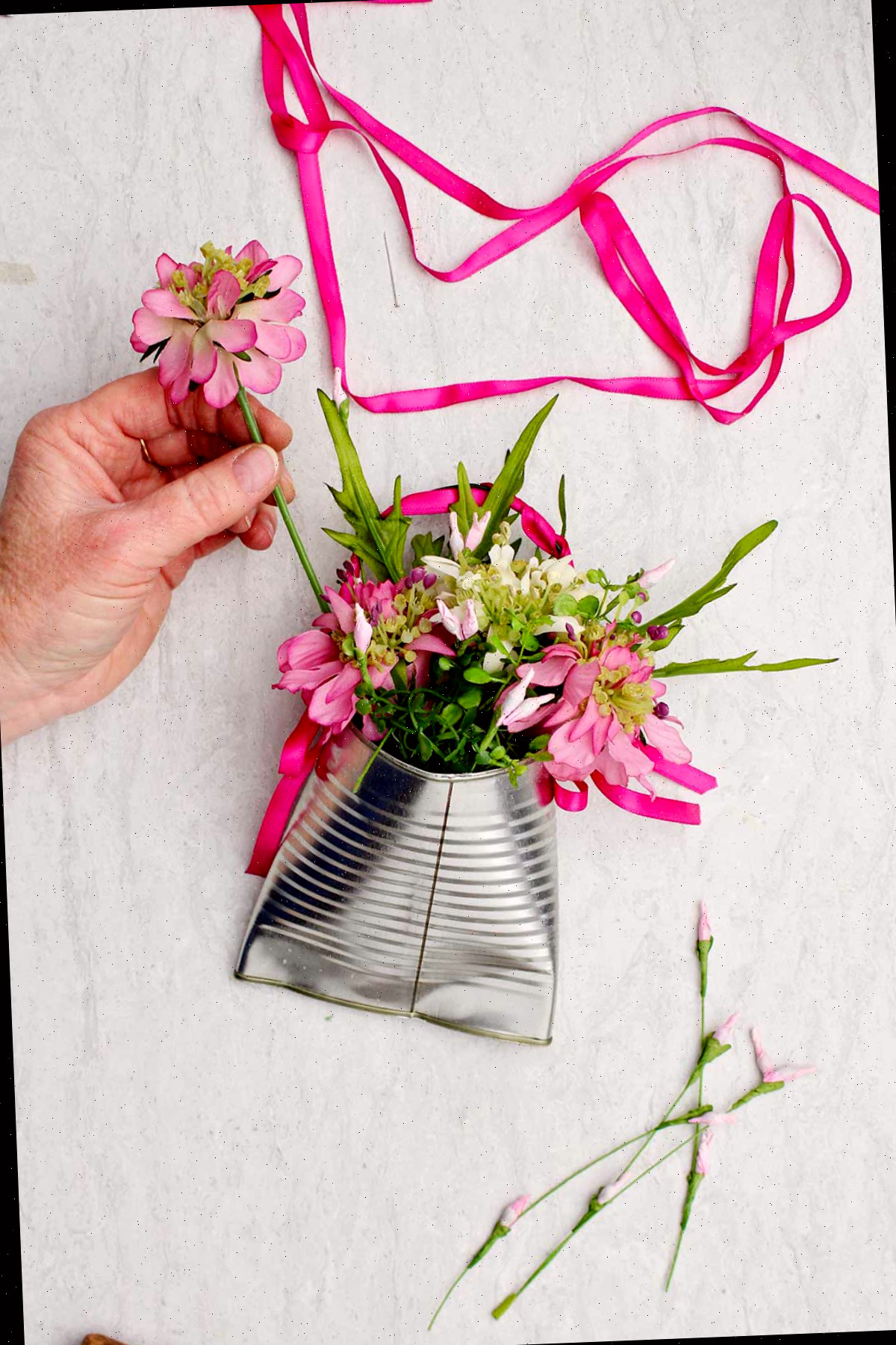 Uma pessoa colocando uma flor rosa em um vaso de lata reciclado completo com fita rosa extra ao lado.