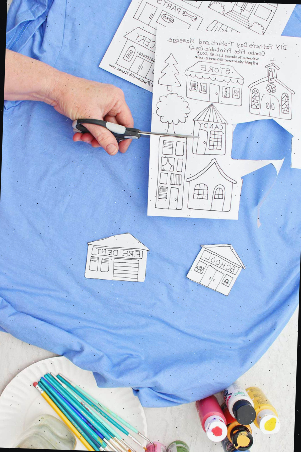 Uma pessoa cortando moldes de diferentes edifícios para a camiseta do Dia dos Pais sobre uma camiseta azul com outros suprimentos próximos.