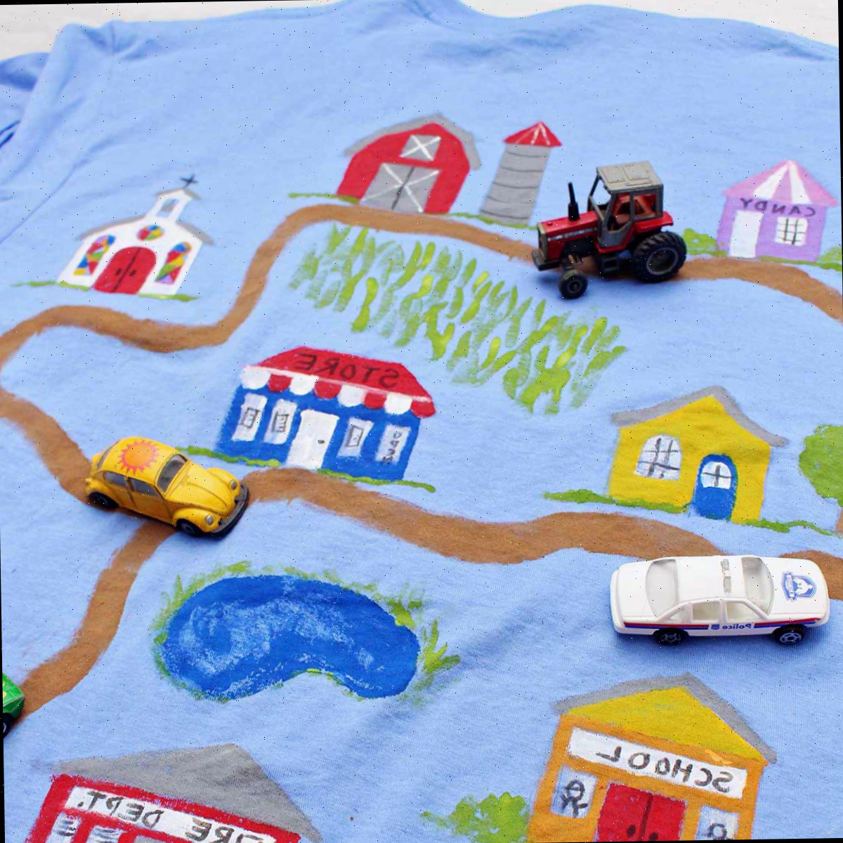 Um carro de polícia, trator e carrinhos de brinquedo Volkswagen Beetle amarelo em uma camiseta do Dia dos Pais pintada com uma estrada de terra levando a vários edifícios.