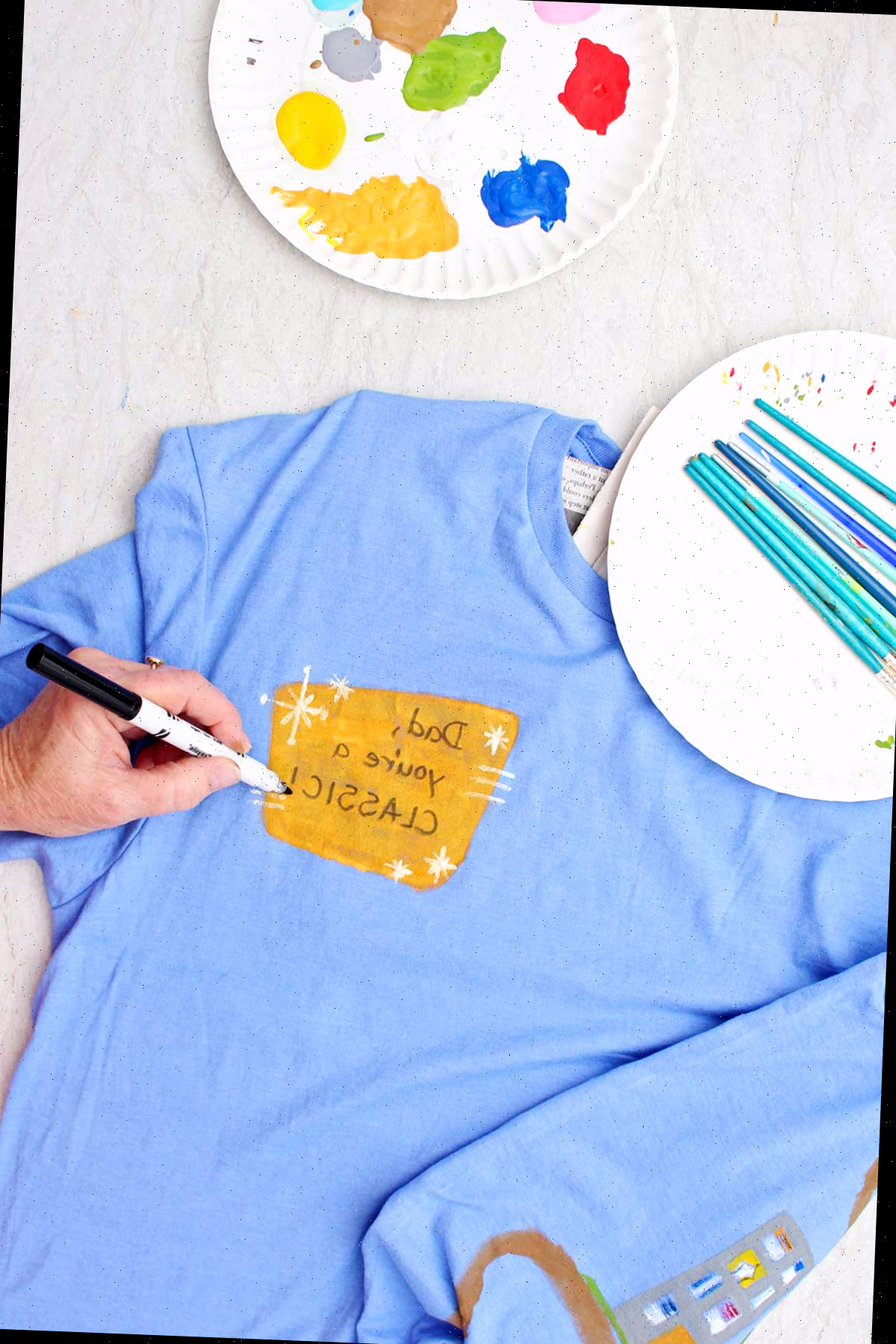 Uma pessoa escrevendo 'Pai, você é um clássico' na parte frontal de uma camiseta azul com um recipiente de tinta e pincéis próximos.