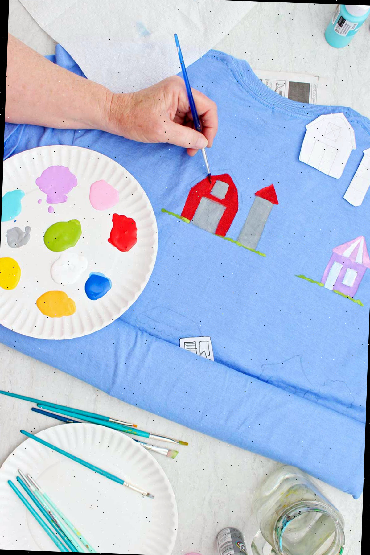 Uma pessoa pintando um celeiro de vermelho em uma camiseta azul com um prato de tinta e outros suprimentos por perto.