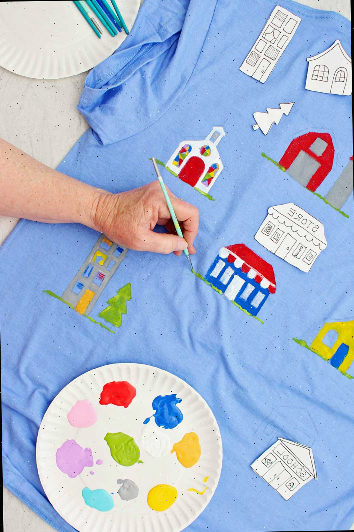 Uma pessoa pintando detalhes de grama perto de uma loja pintada em uma camiseta azul com um prato de tinta por perto.