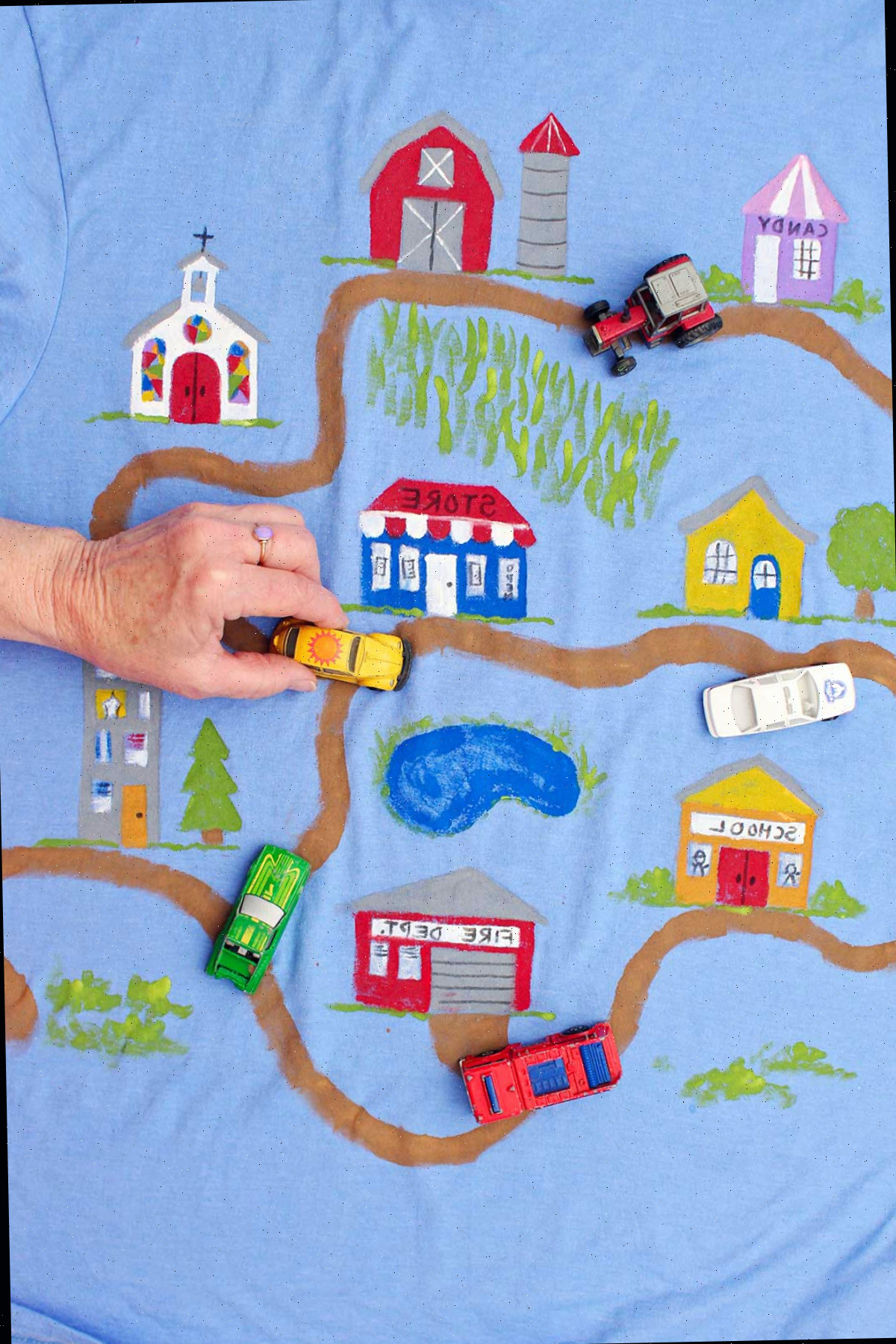 Pessoa movimentando um carro de brinquedo Volkswagen Beetle amarelo em uma camiseta do Dia dos Pais decorada com uma estrada de terra levando a diversos edifícios.