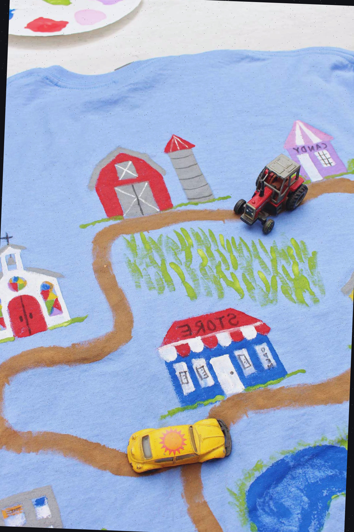 Um trator e um carro de brinquedo Volkswagen Beetle amarelo em uma camiseta do Dia dos Pais decorada com uma estrada de terra levando a vários edifícios, com um recipiente de tinta ao fundo.