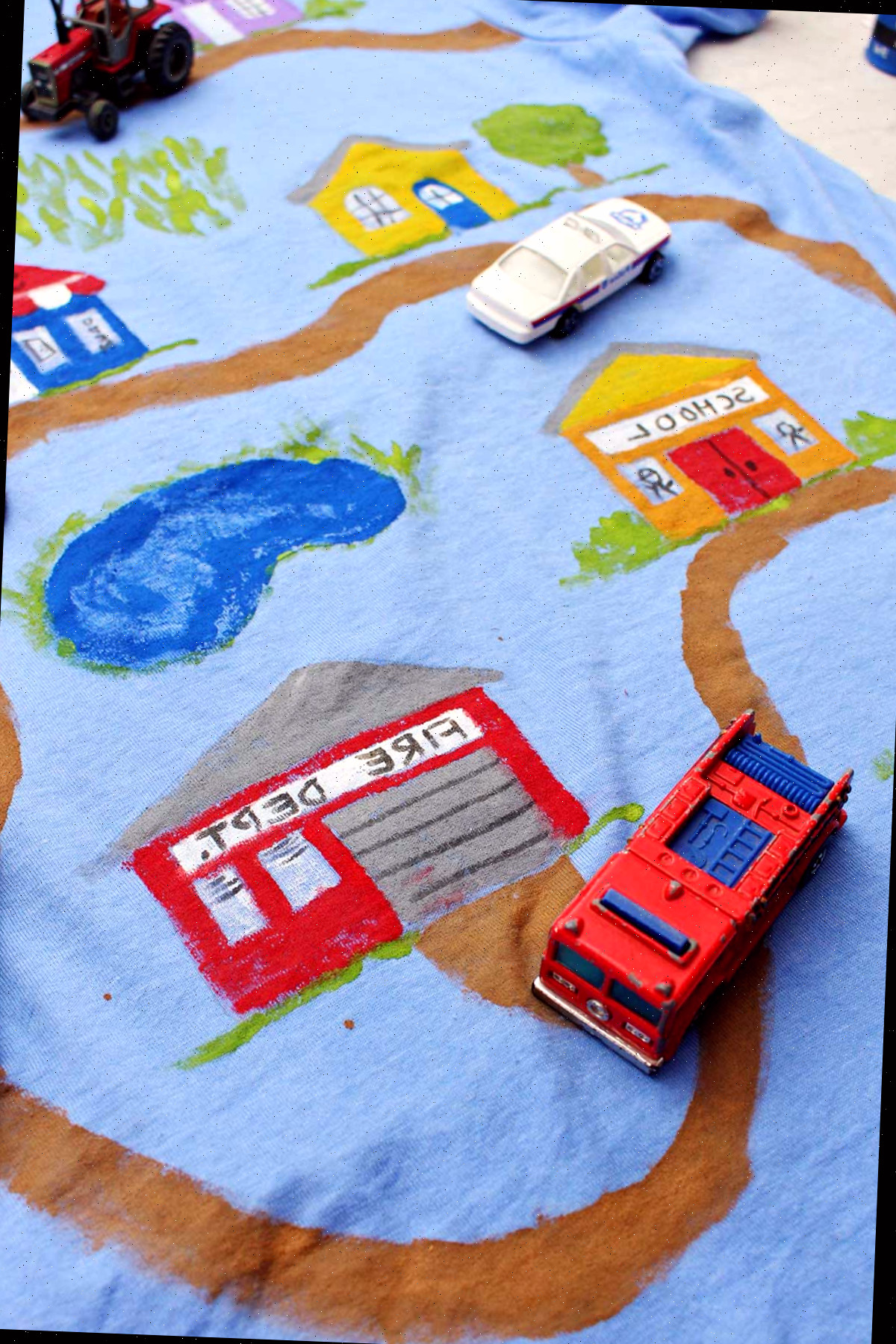 Um carro de polícia, trator e caminhão de bombeiros de brinquedo sobre uma camiseta do Dia dos Pais decorada com uma estrada de terra levando a vários edifícios.