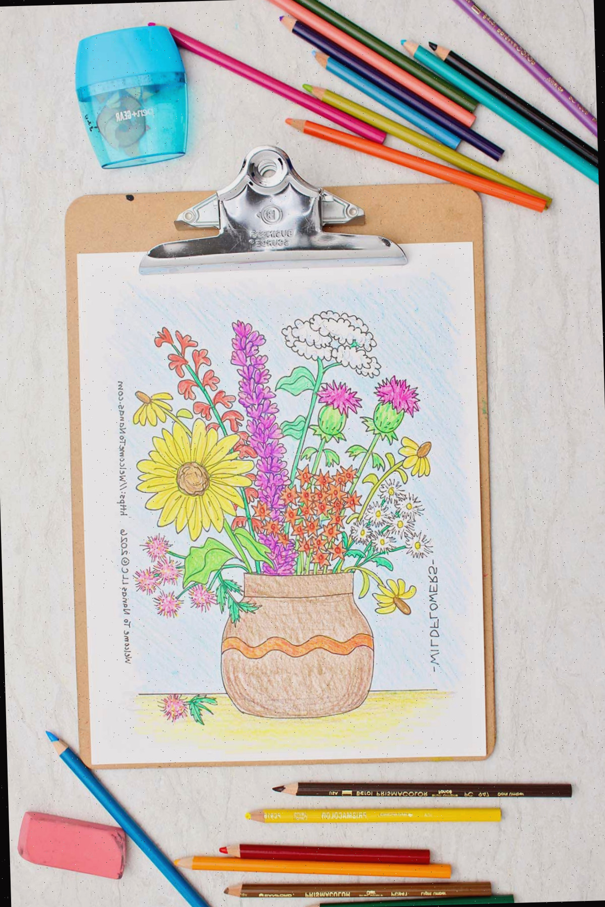Uma página para colorir de um vaso marrom repleto de flores silvestres coloridas preso a uma prancheta com lápis de cor espalhados ao redor.