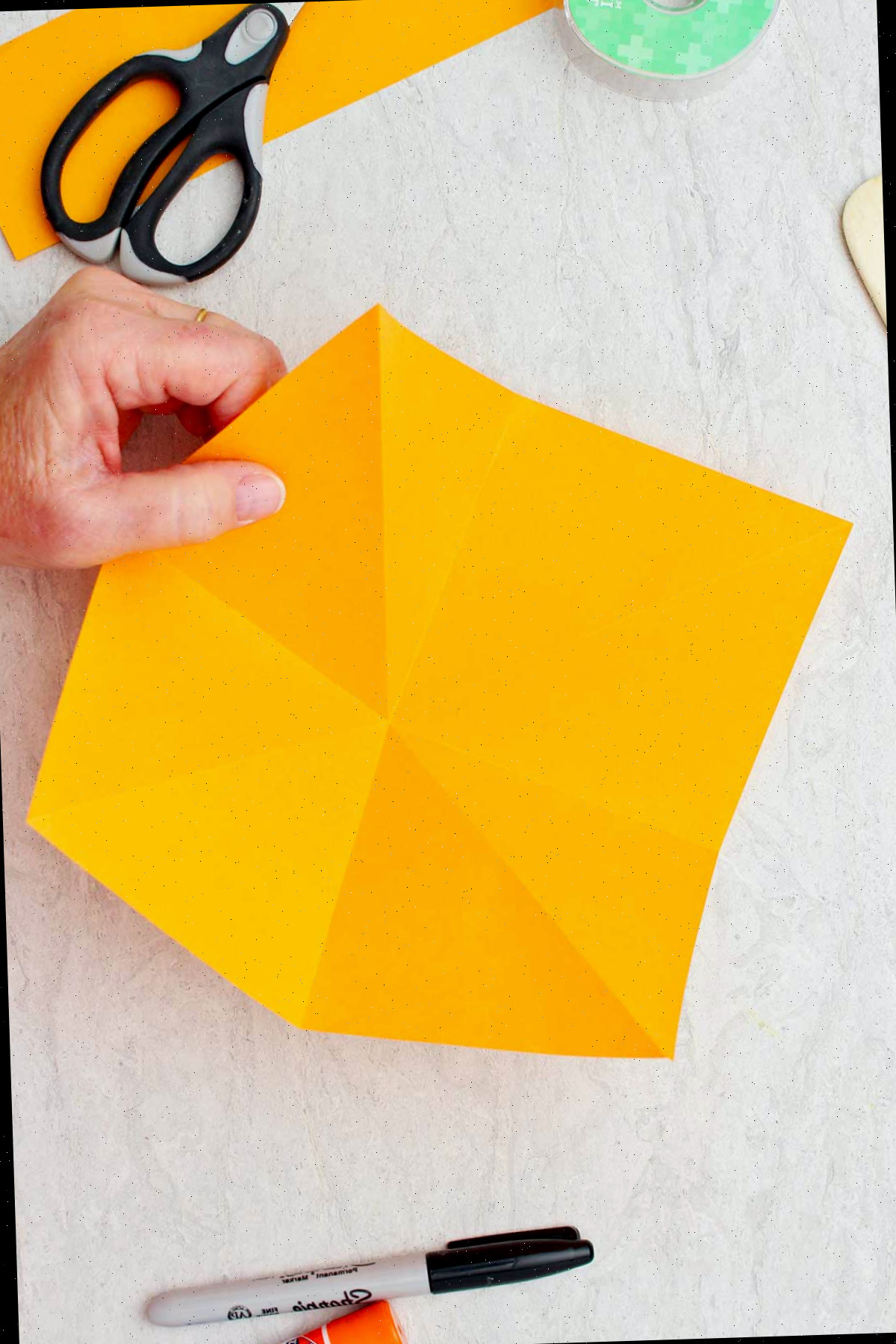 Mão segurando o lado direito de um pedaço cortado de papel laranja pronto para ser dobrado em uma abóbora de origami.