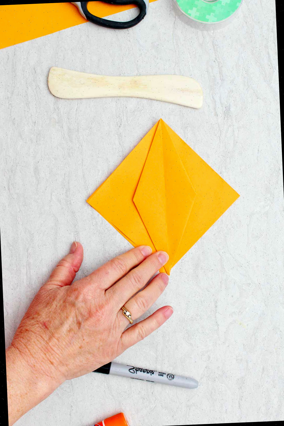 Um pedaço quadrado de papel laranja com um diamante dobrado no centro e outros materiais ao redor para fazer abóboras de origami.