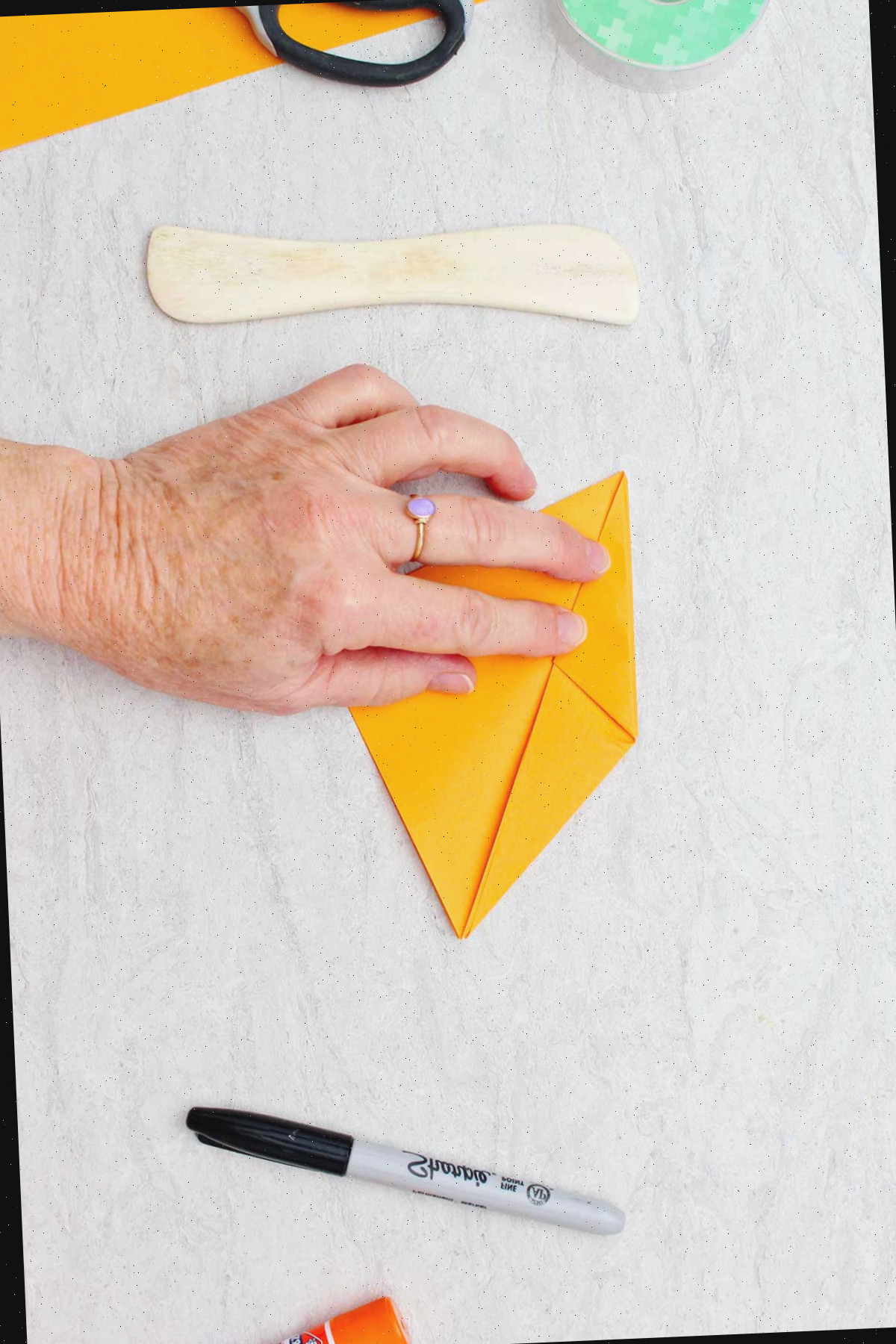 Indivíduo dobrando um pedaço de papel laranja para confeccionar uma abóbora de origami com outros materiais ao redor.