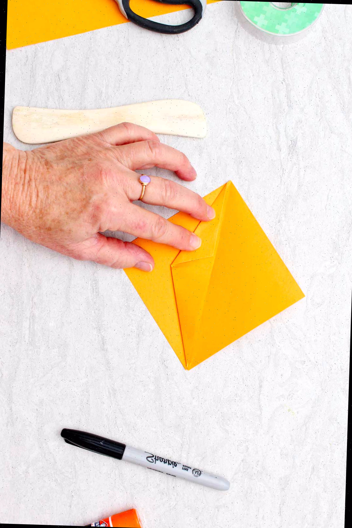Indivíduo dobrando um pedaço de papel laranja para fazer uma abóbora de origami com outros materiais ao redor.