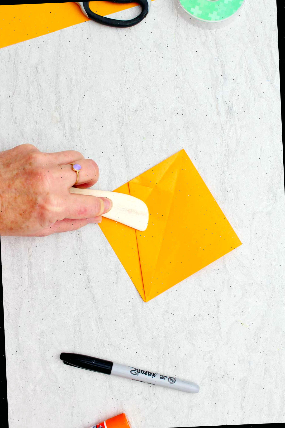 Pessoa usando um bastão de madeira arredondado para afinar as bordas dobradas de um pedaço de papel laranja utilizado para criar uma abóbora de origami.