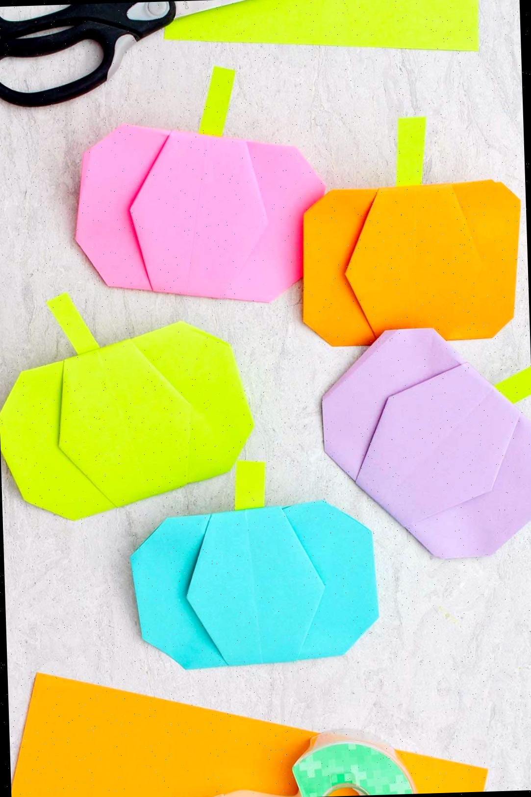 Cinco abóboras de origami de diferentes cores com tesouras, fita e outros pedaços de papel colorido ao redor.