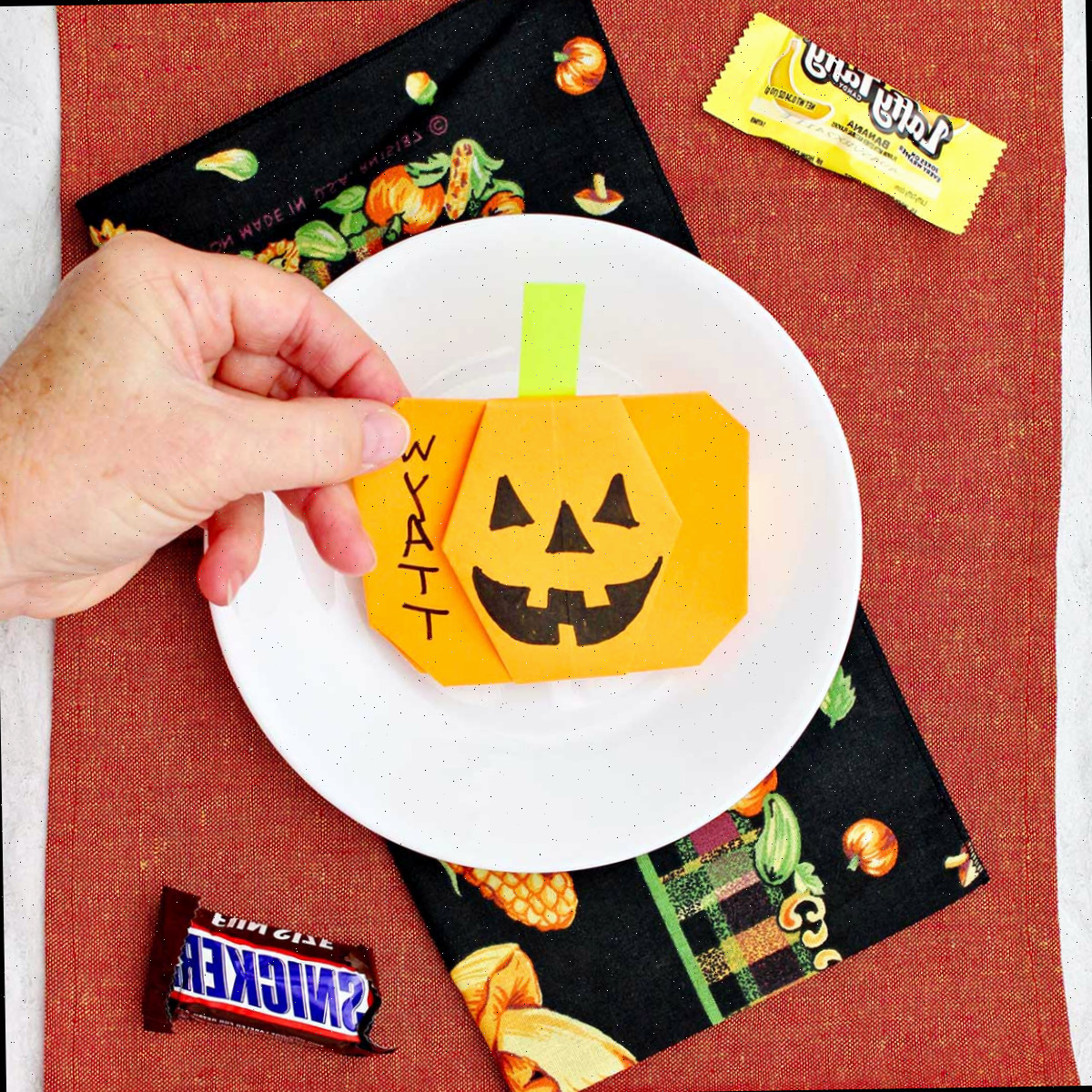 Mão posicionando uma abóbora de origami laranja em um lugar para uma configuração com alguns doces de Halloween ao redor.