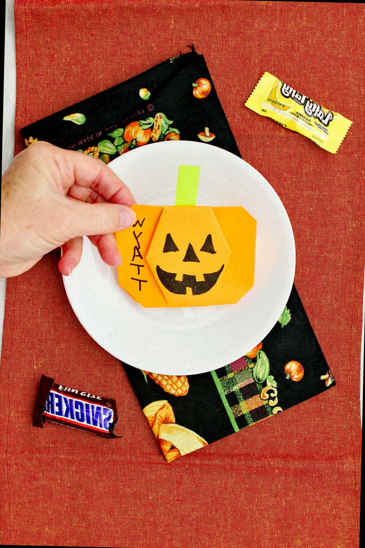 Mão colocando uma abóbora de origami laranja em um lugar para uma configuração de mesa com alguns doces de Halloween ao redor.