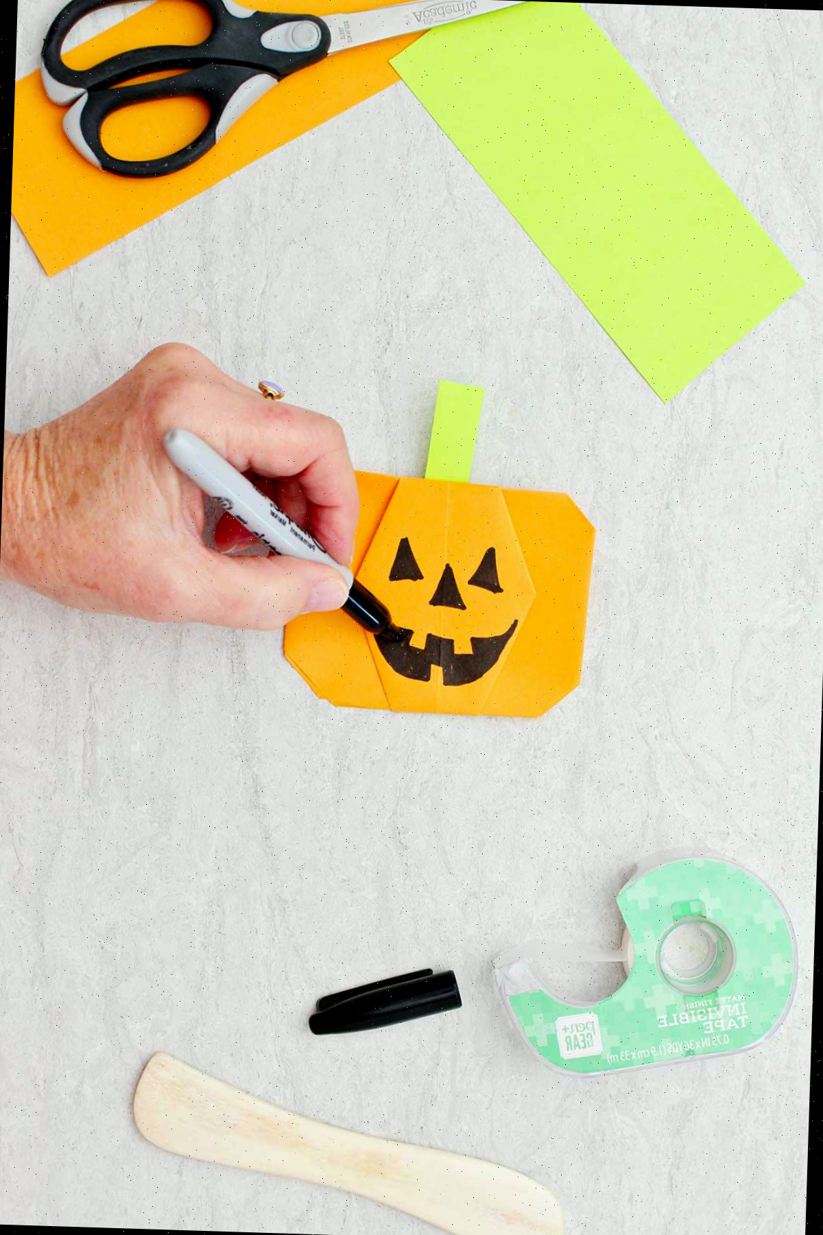 Uma pessoa desenhando um rosto de jack-o-lantern em uma abóbora de origami laranja com outros suprimentos ao redor.