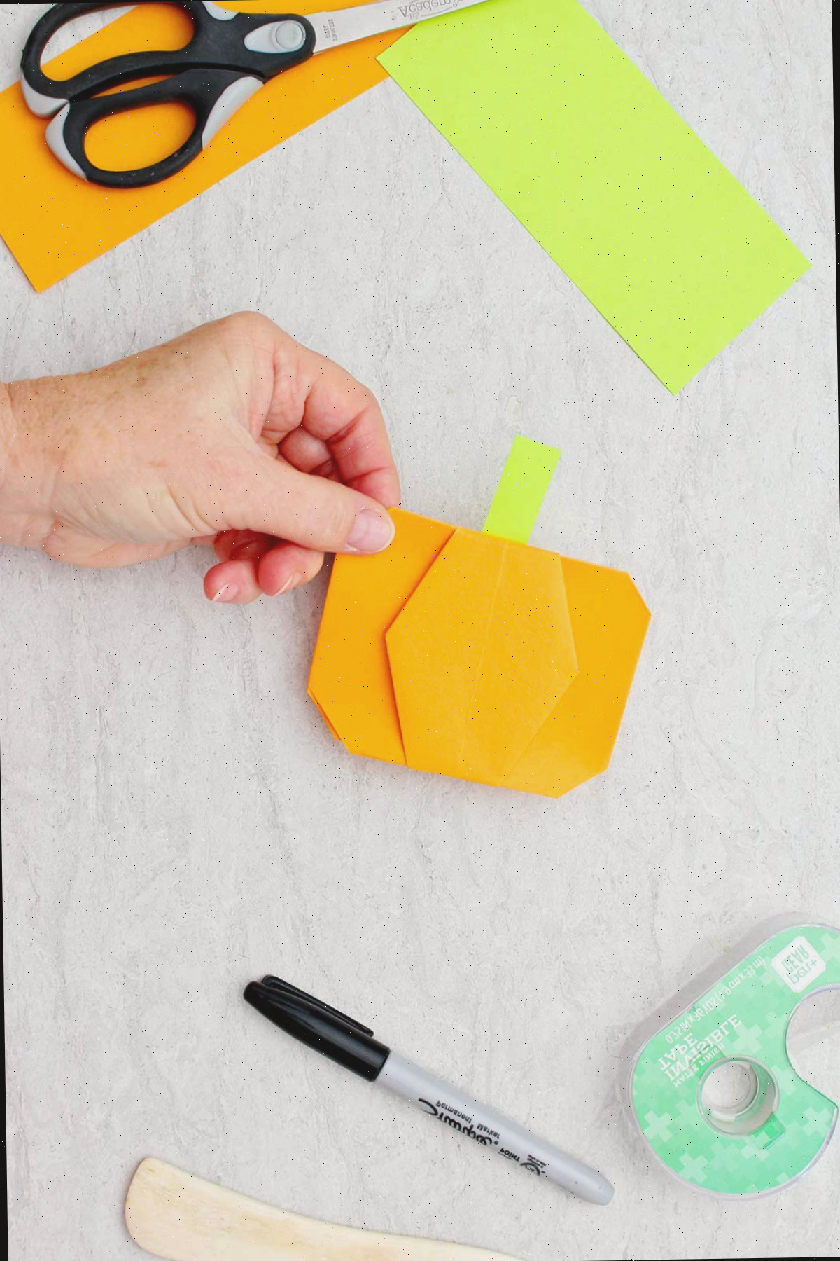 Pessoa segurando uma abóbora de origami laranja completa com um talo verde-lima rodeada por outros materiais para o projeto de abóbora de origami.