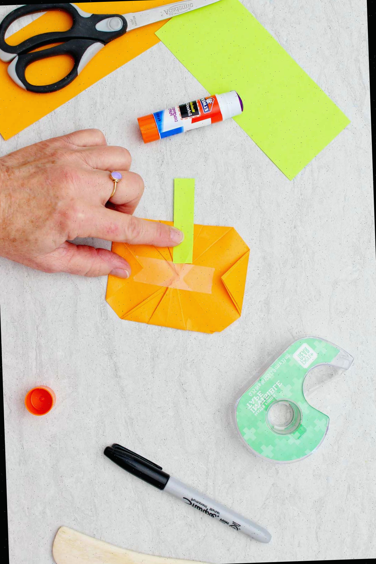 Uma pessoa aplicando um retângulo verde na parte de trás de uma abóbora de origami laranja como um talo, cercada por outros materiais para criar abóboras de origami.