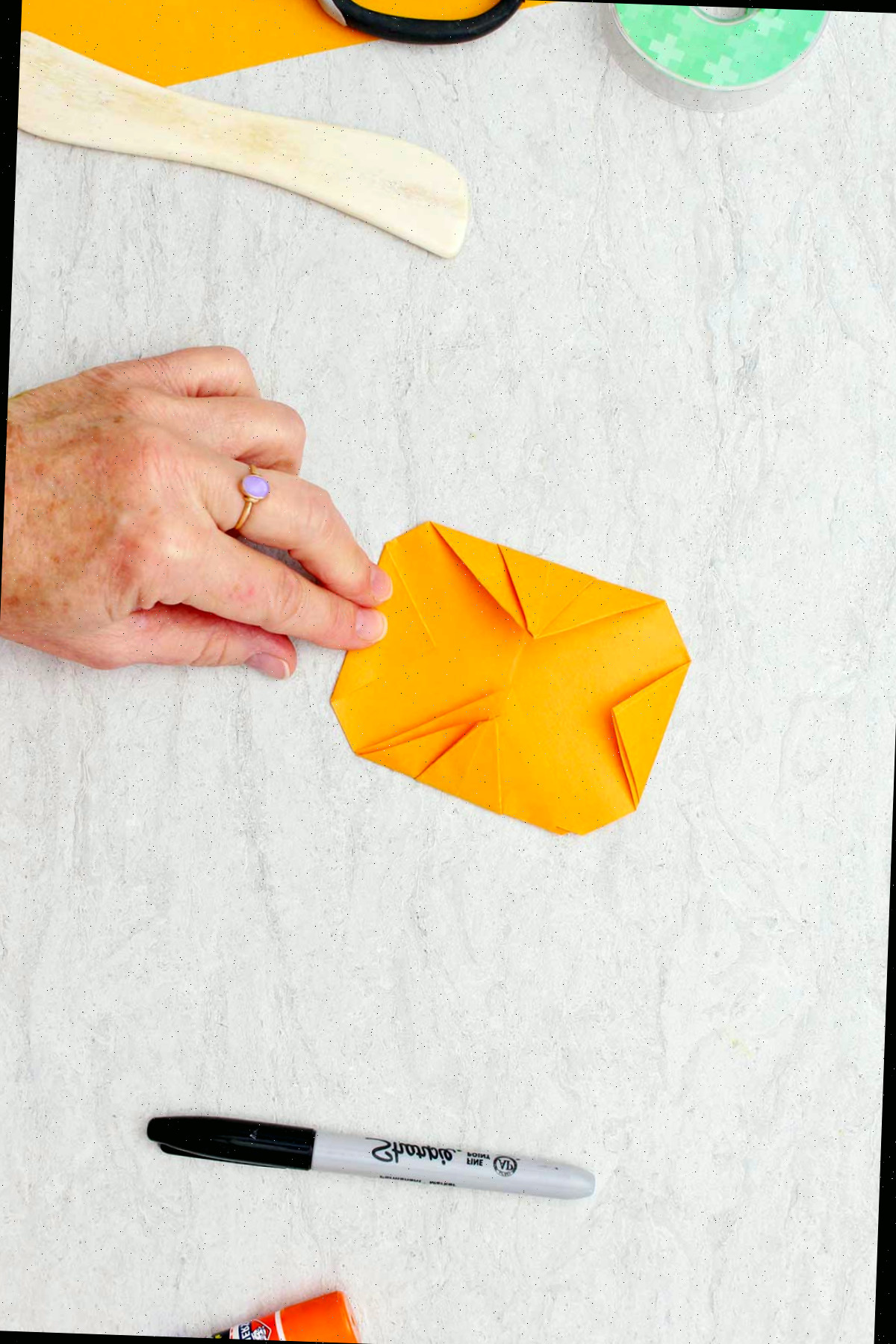 Indivíduo dobrando uma extremidade de papel laranja e mostrando o lado reverso de uma abóbora de origami quase completada.