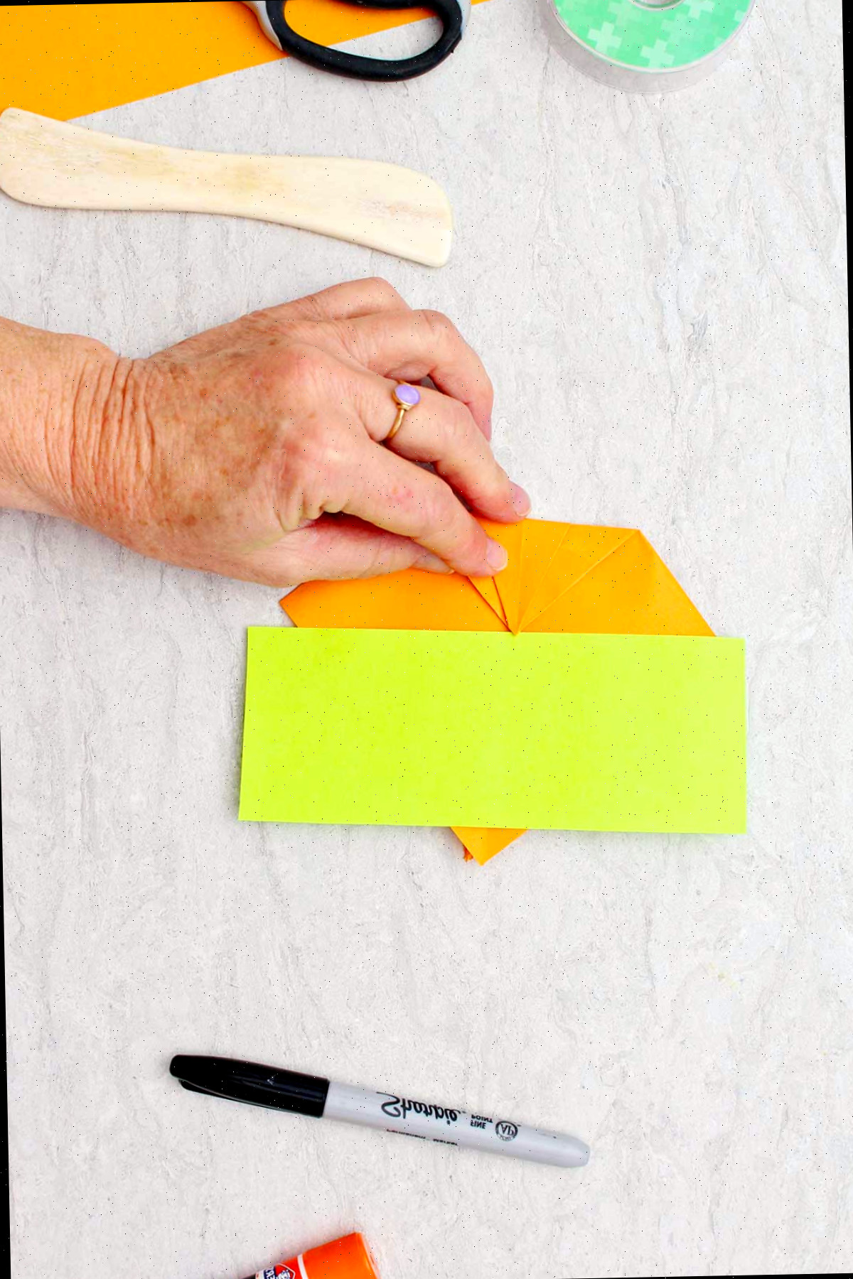 Um retângulo verde de papel repousando sobre um pedaço de papel laranja que uma pessoa está dobrando, com outros materiais nas proximidades para fazer abóboras de origami.