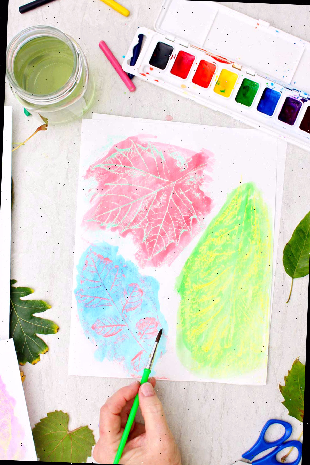 Uma pessoa pintando com aquarela sobre impressões de folhas com folhas e aquarelas espalhadas ao redor.