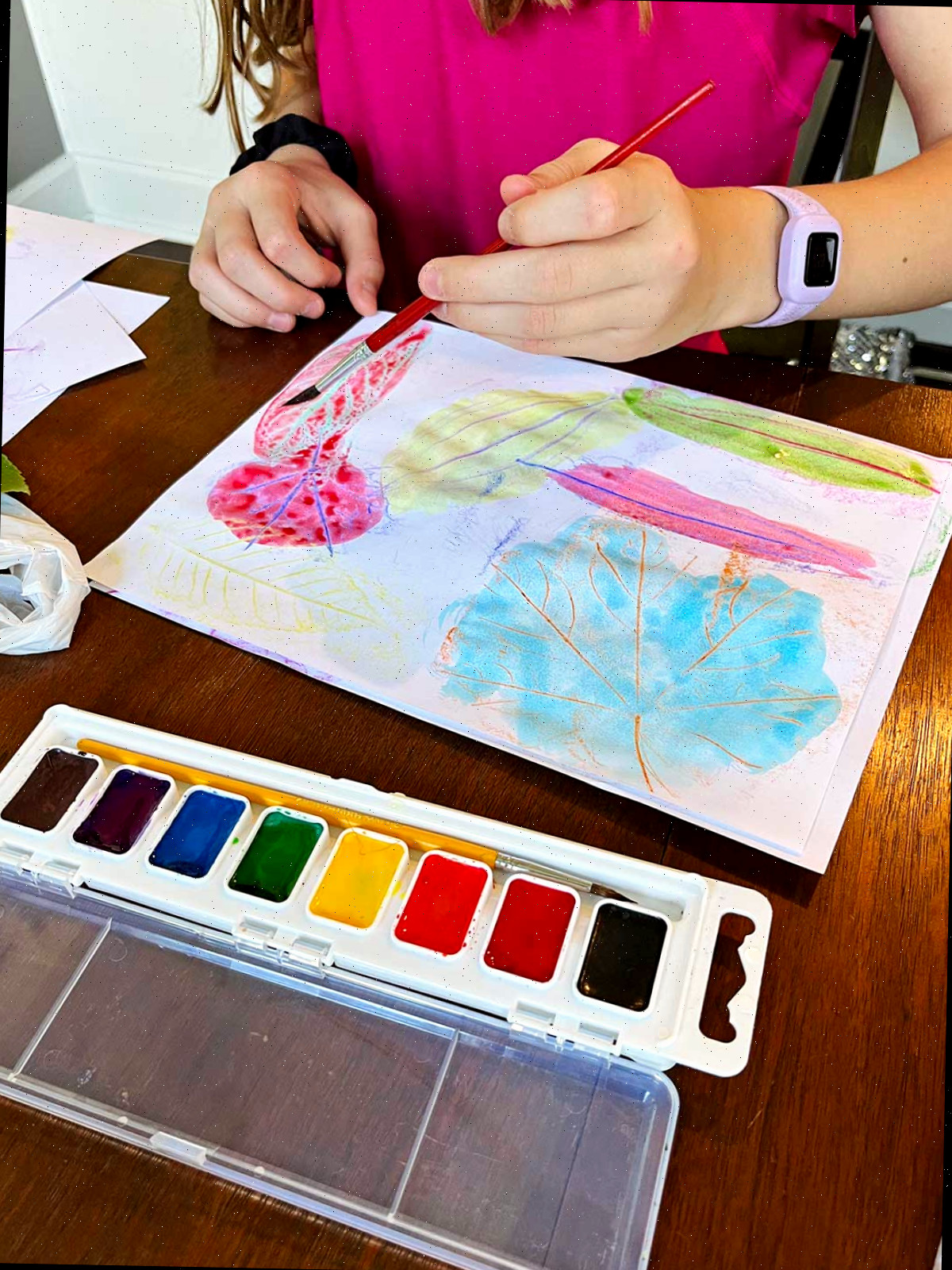 Uma menina jovem com uma camisa rosa pintando com aquarela sobre impressões de folhas em uma mesa.