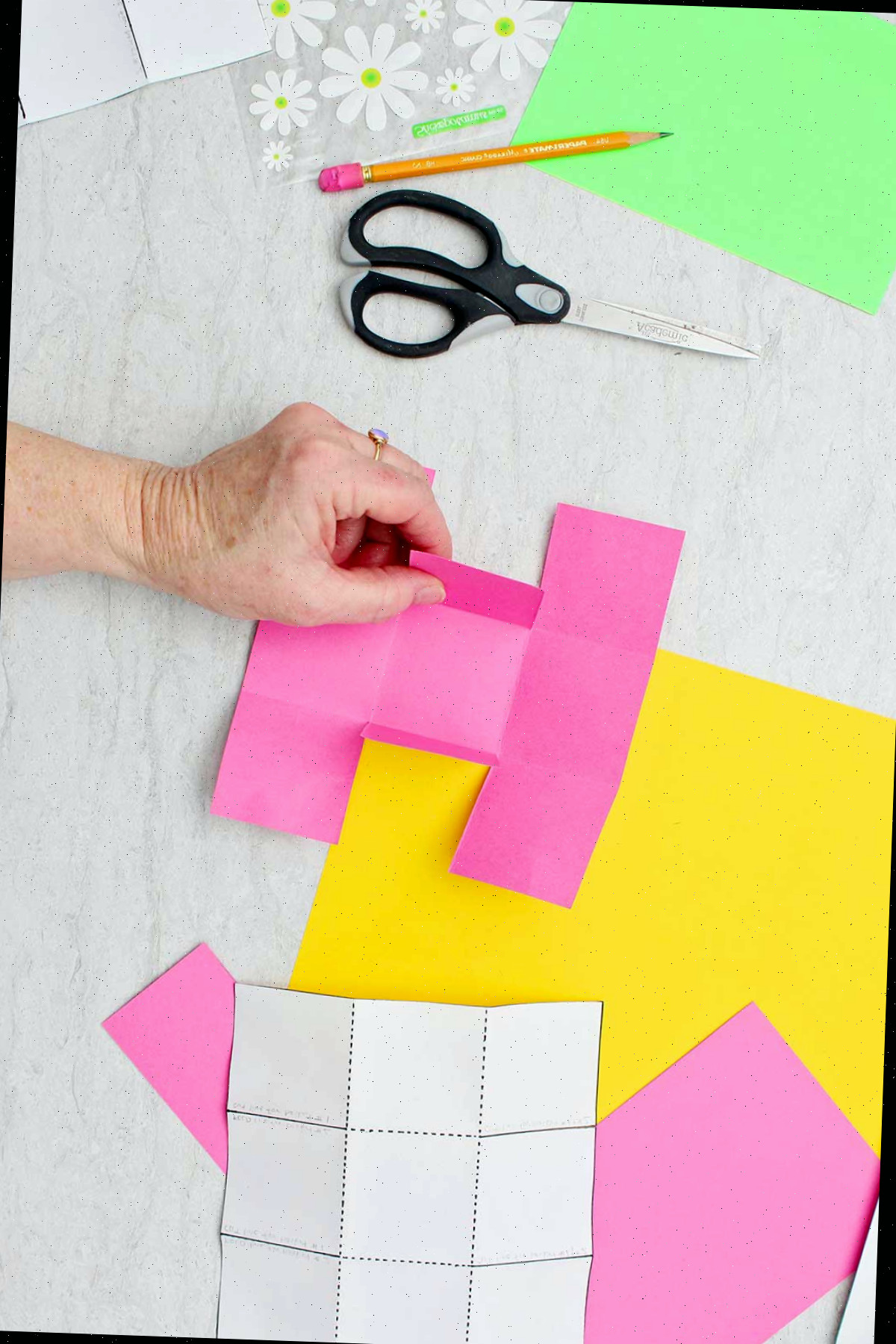 Indivíduo dobrando o quadrado central em direção ao centro de um grande quadrado rosa de papel, quase assemelhando-se a uma letra "H".