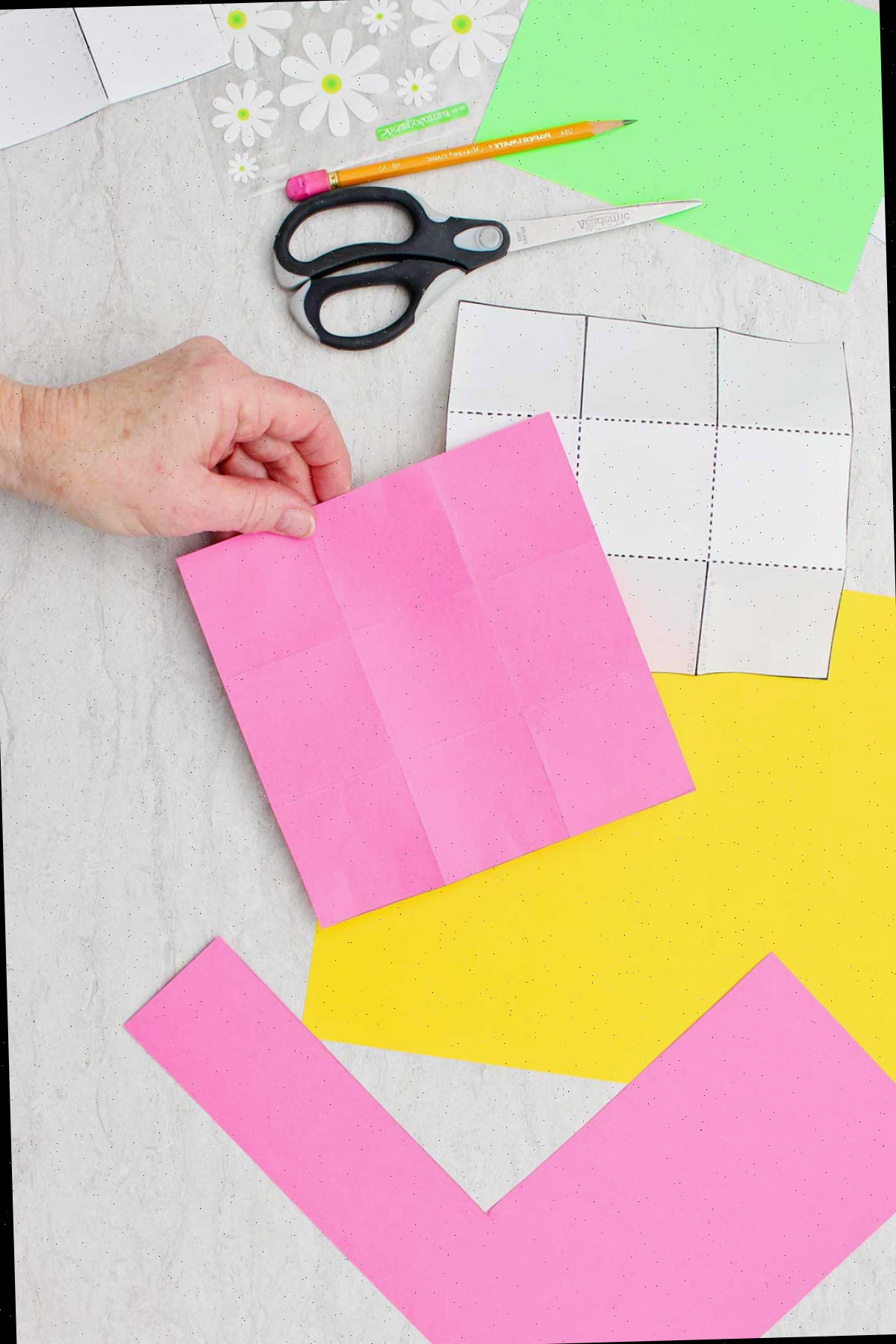 Uma pessoa segurando um pedaço quadrado de papel rosa com linhas de dobra para criar nove caixas iguais com outros suprimentos ao redor.