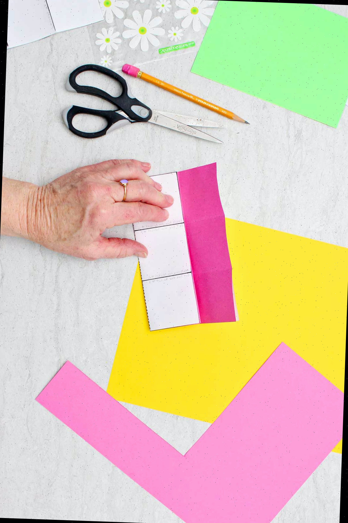 Pessoa segurando um molde recortado próximo a um pedaço de papel rosa escuro para verificar linhas de dobra com outros materiais próximos.