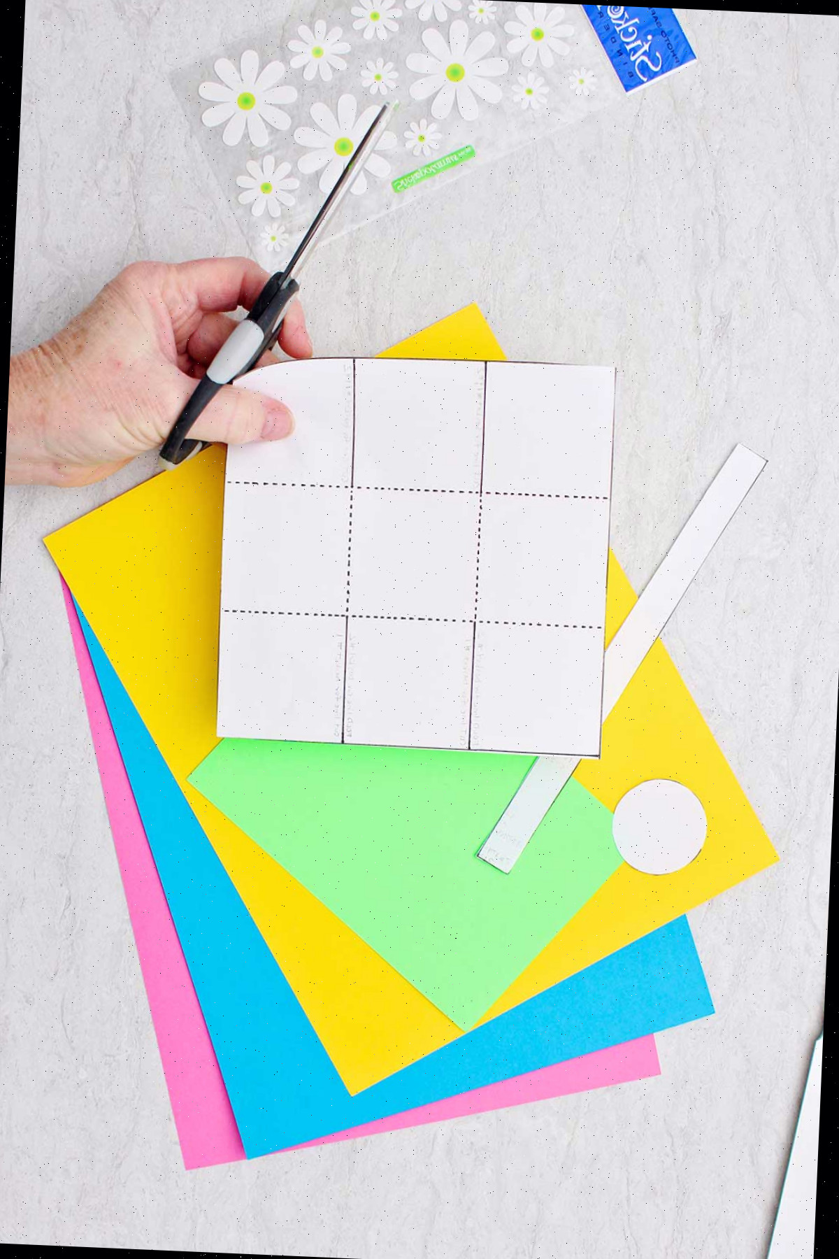 Pessoa segurando um molde para um mini cesto de Páscoa sobre uma pilha de papel colorido com uma folha de adesivos de flores próxima.