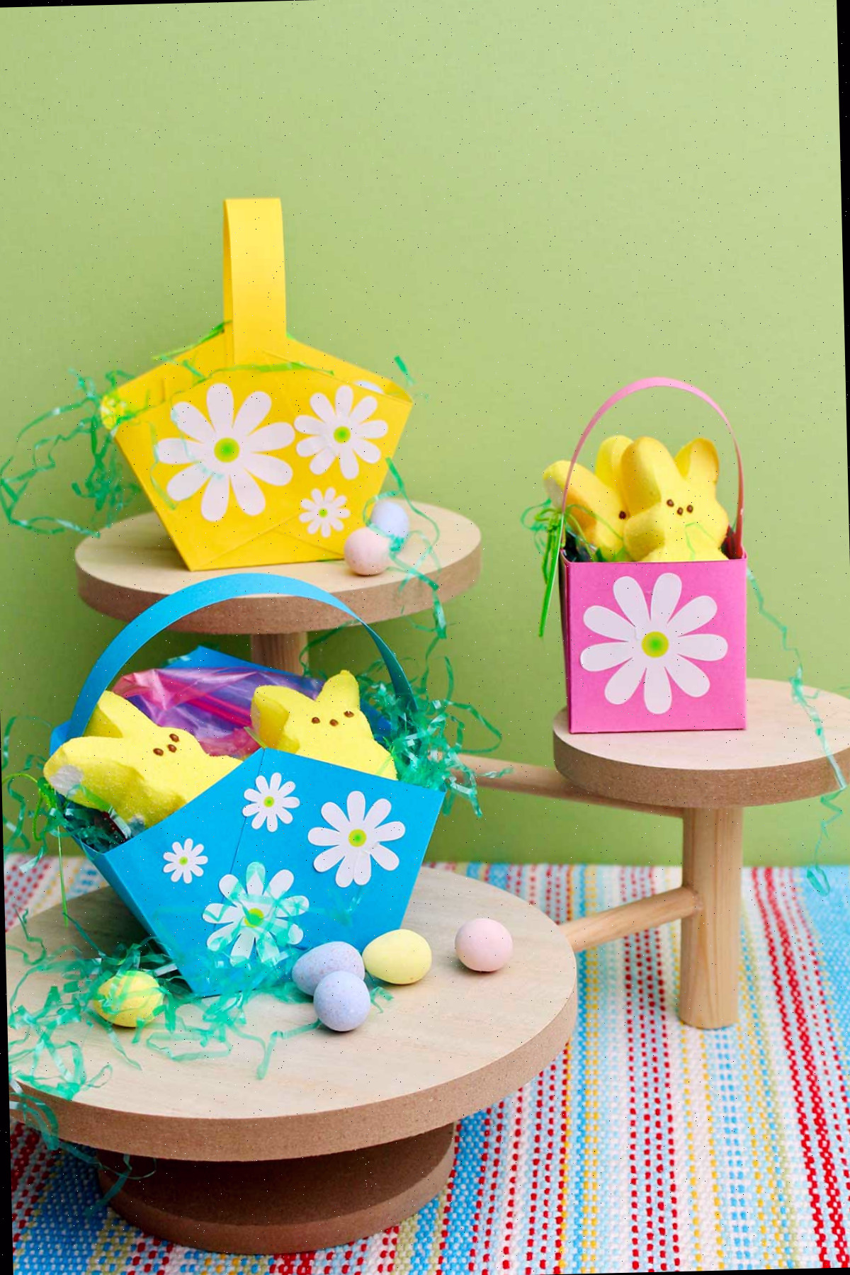 Três mini cestas de Páscoa repletas de doces com flores brancas decoradas em pedestais de madeira contra um fundo verde.