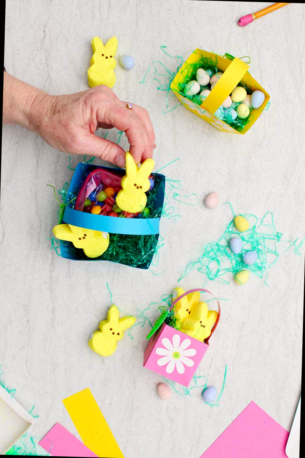 Indivíduo colocando um Coelhinho de Marshmallow amarelo em uma mini cesta de Páscoa azul repleta de doces e grama de Páscoa, com outras mini cestas de Páscoa já finalizadas ao lado.