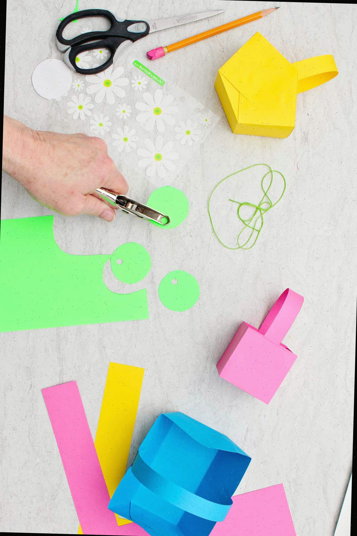 Indivíduo perfurando buracos em círculos cortados de papel verde para confeccionar etiquetas para as mini cestas de Páscoa.