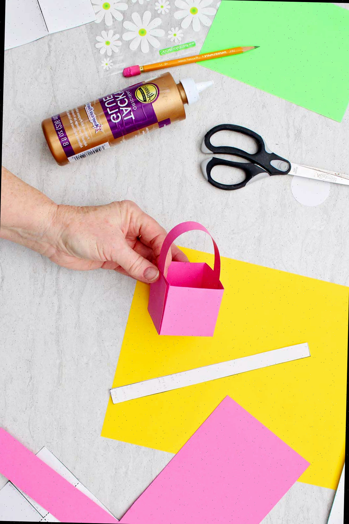 Indivíduo fixando a alça em um mini cesto de Páscoa de papel rosa com outros materiais ao redor.
