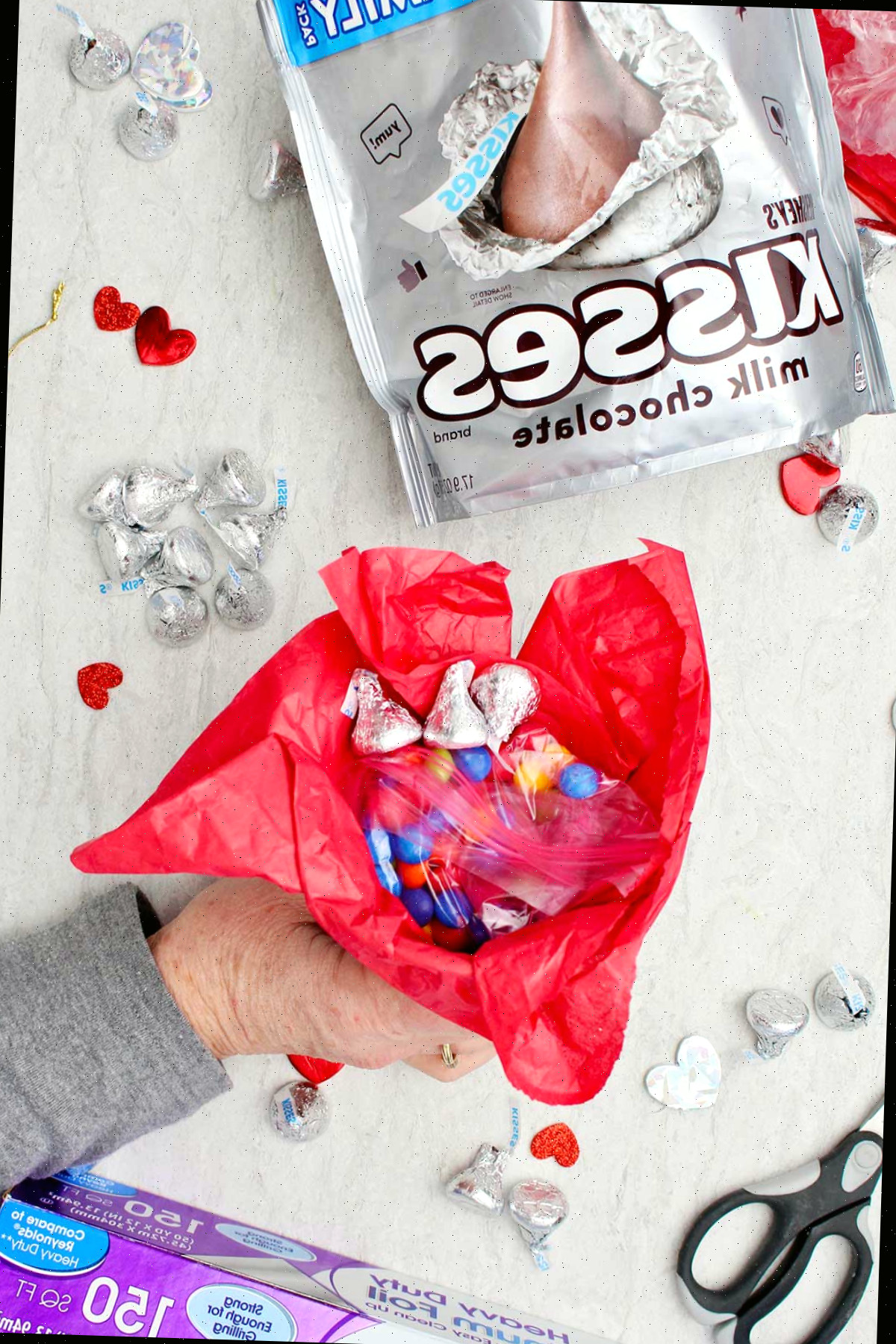 Pessoa segurando um funil com papel de seda vermelha e guloseimas do Dia dos Namorados dentro com outros suprimentos ao redor.