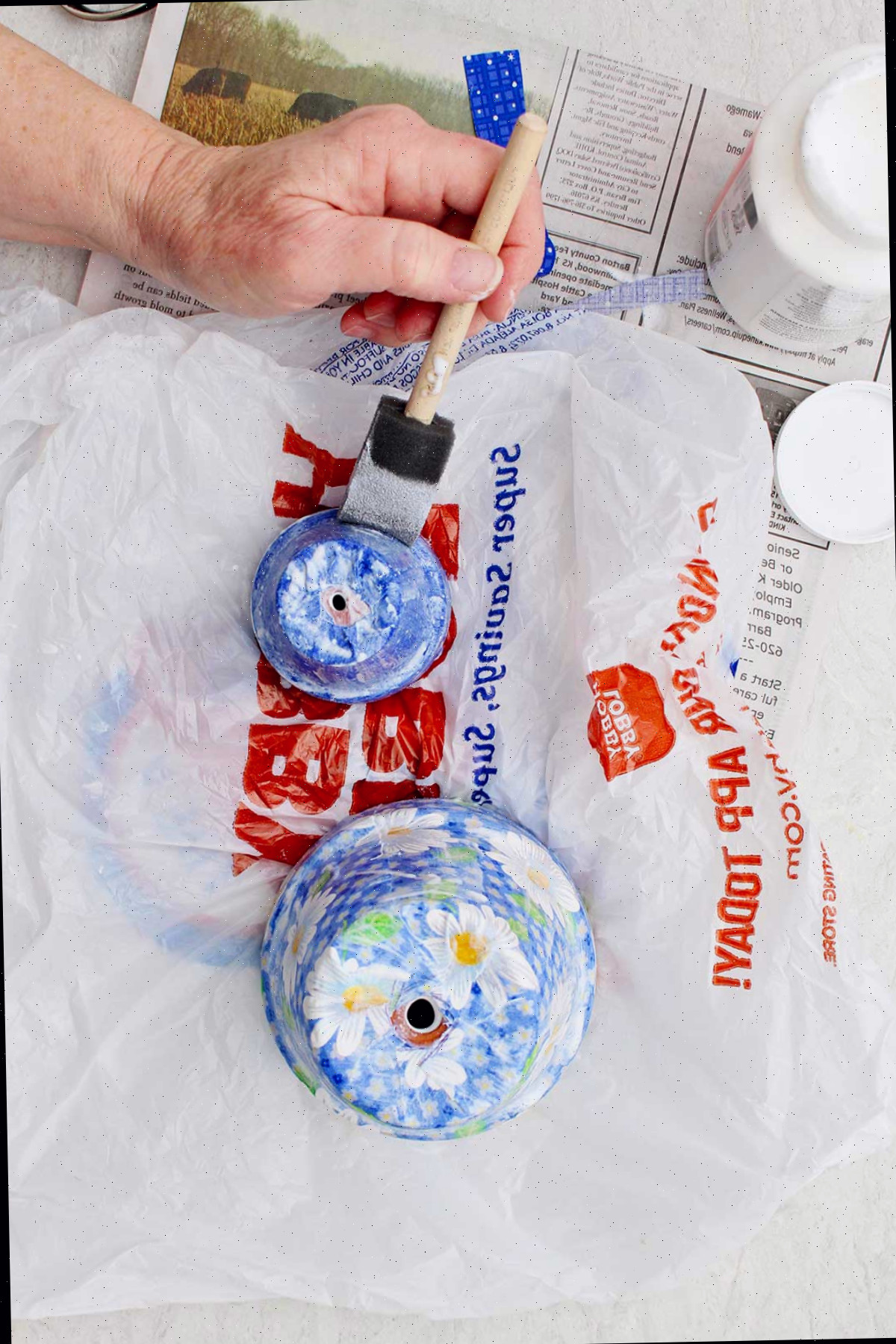 Uma pessoa aplicando mod podge em um vaso pequeno com outro vaso maior secando ao lado.