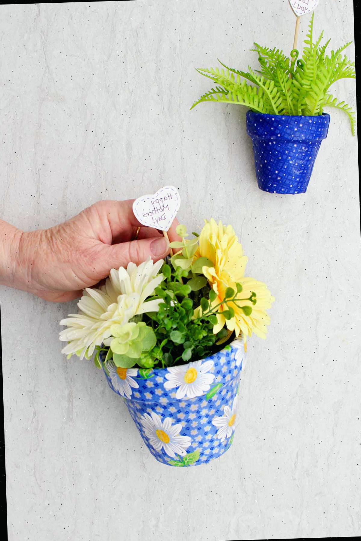 Uma pessoa colocando um post em forma de coração dizendo "Feliz Dia das Mães" dentro de um vaso de flores decoupado com flores.