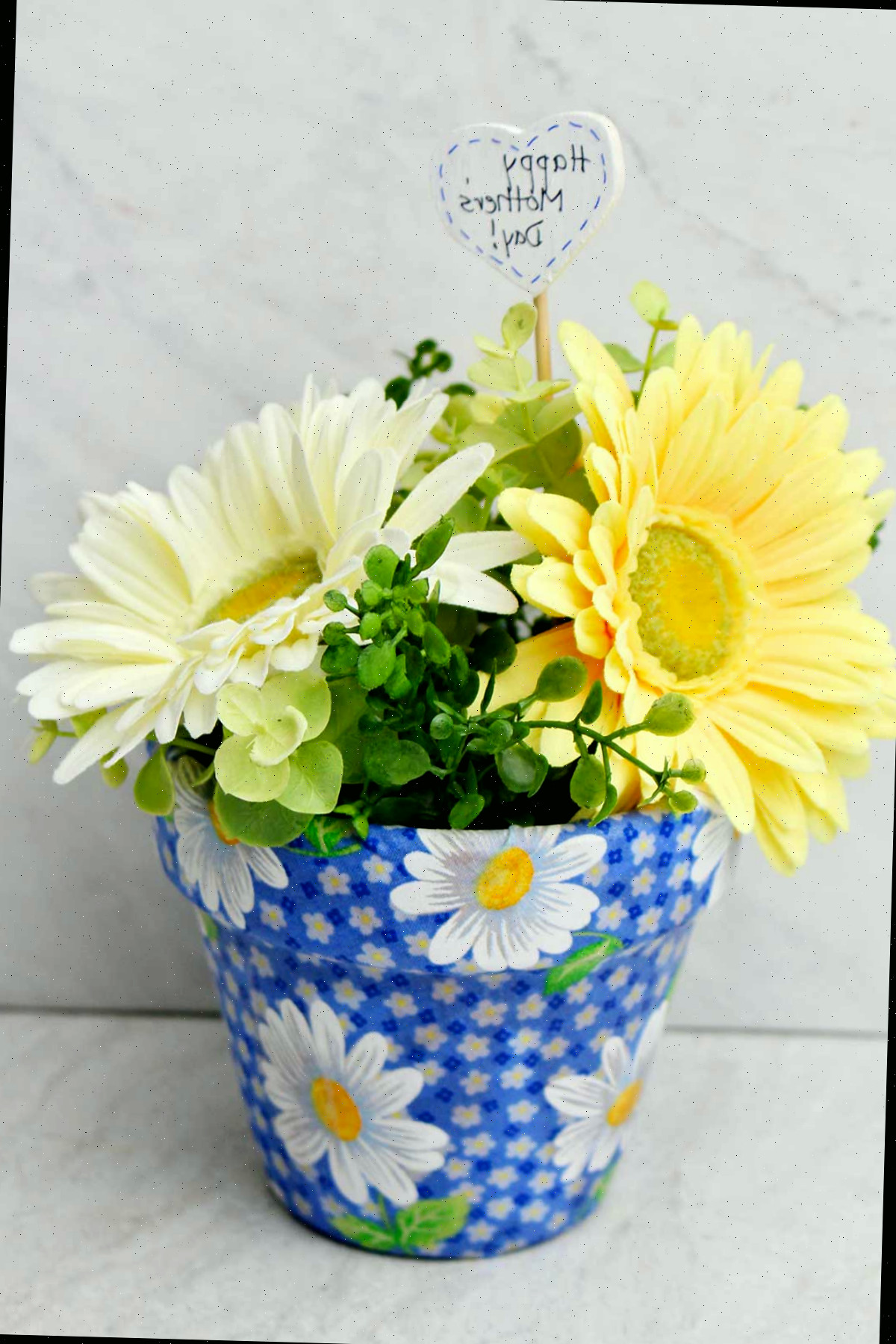 Um vaso decoupado com tecido azul de margaridas, com flores de primavera dentro e um post em forma de coração dizendo "Feliz Dia das Mães"