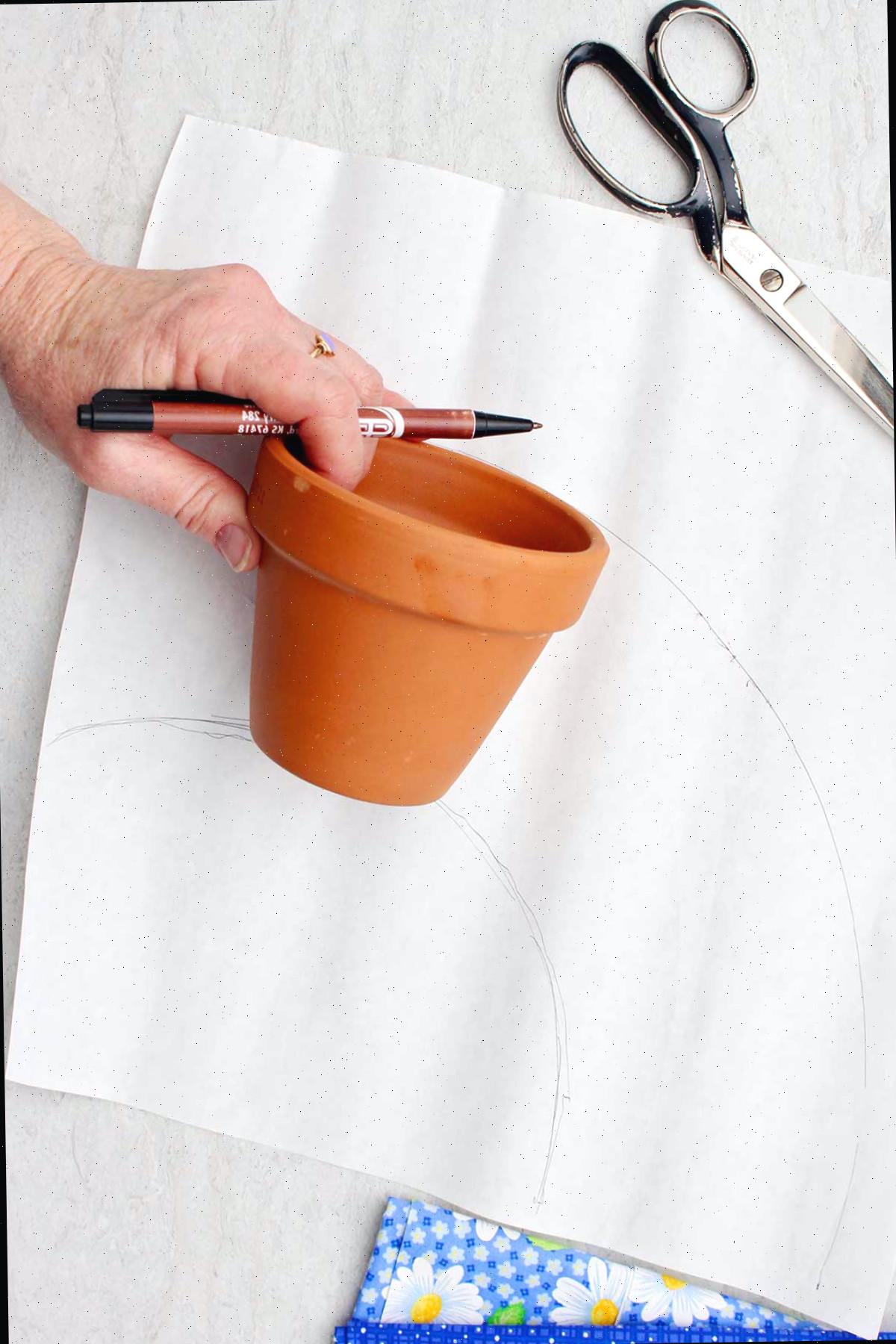 Uma pessoa traçando ao redor de um vaso de flores para ver quanto tecido ela precisará para cobri-lo.