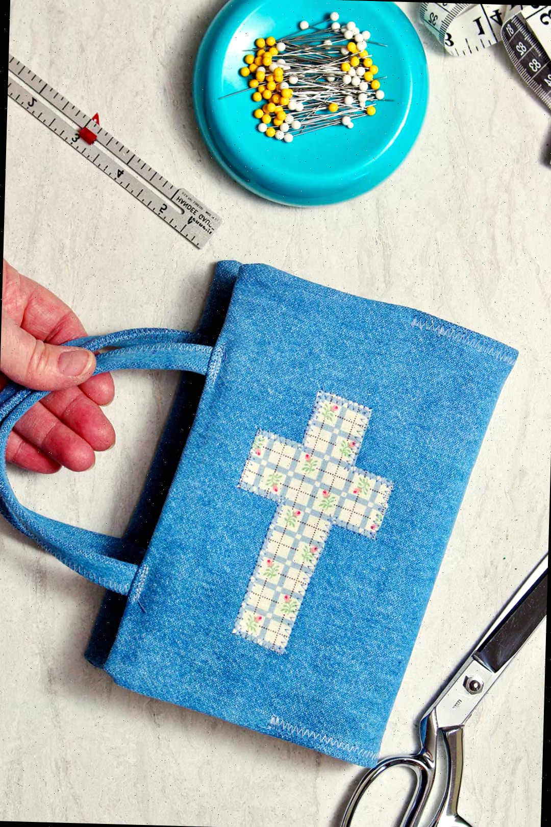 Uma capa de livro de tecido finalizada com alças. O livro é feito de tecido de denim e possui uma pequena cruz na frente.