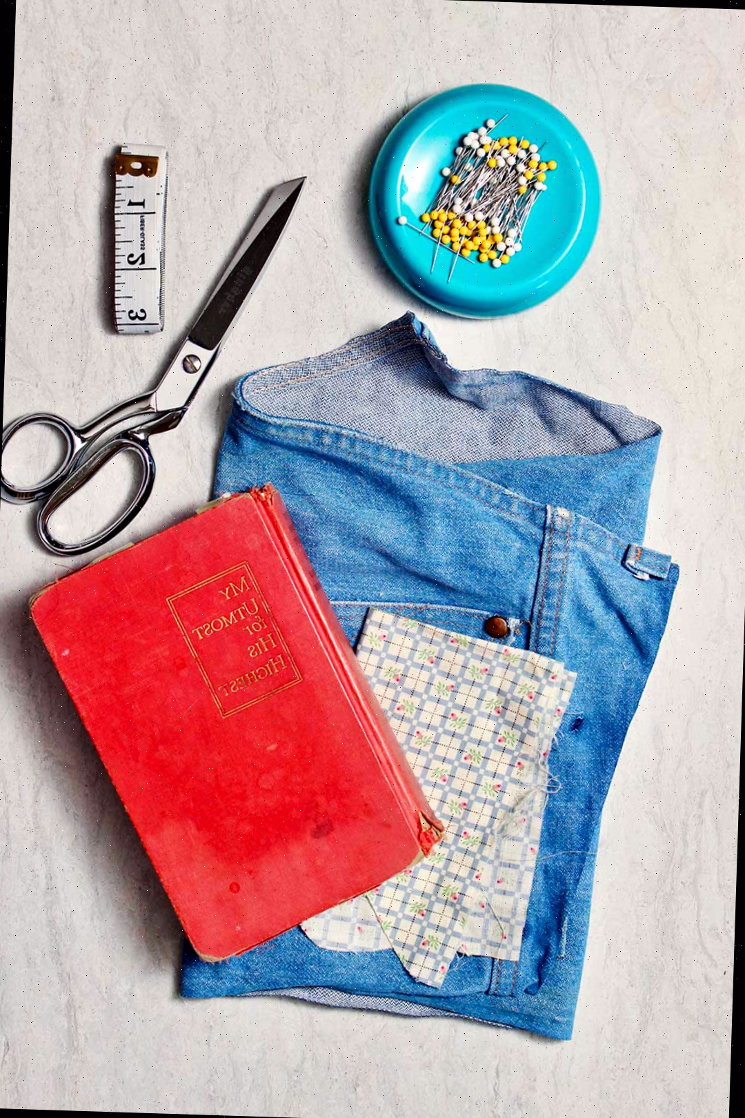 Materiais necessários para confeccionar uma capa de livro de tecido. Retalhos de calças de denim velhas, um livro, alfinetes, tesouras e uma fita métrica.