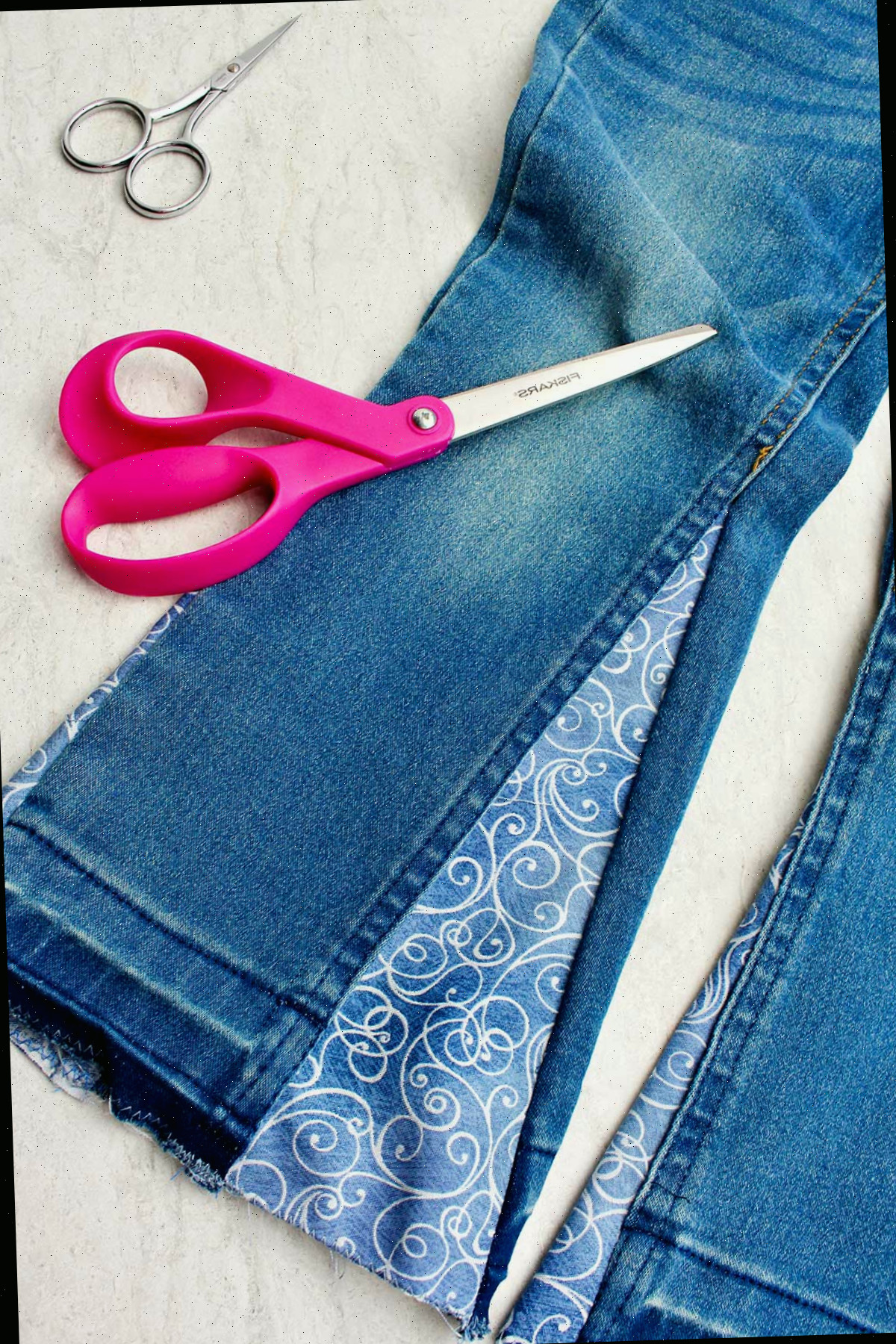 Um par de jeans do lado certo, com tecido decorativo costurado na lateral para fazer barra sino, com dois pares de tesouras por perto.