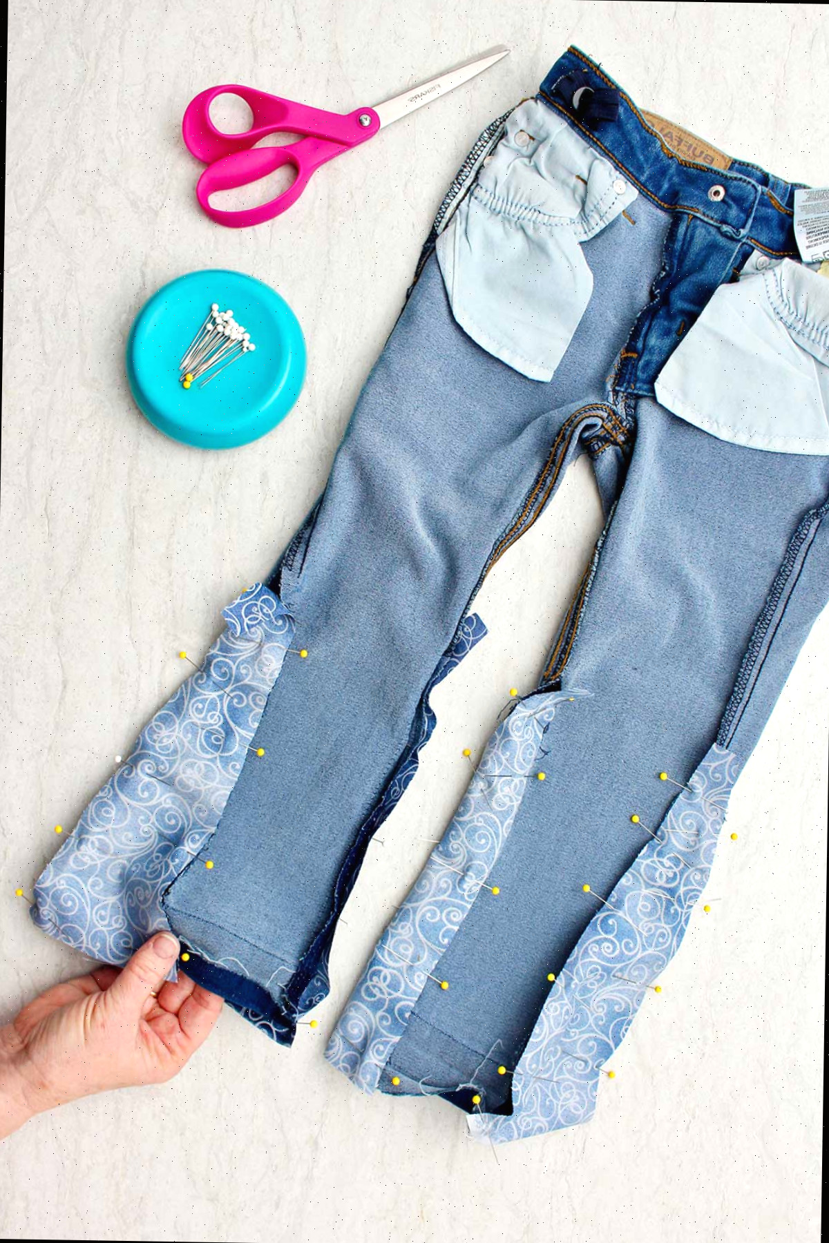 Jeans do avesso com alfinetes segurando o tecido no lugar para fazer barra de sino, com alfinetes e tesouras por perto.