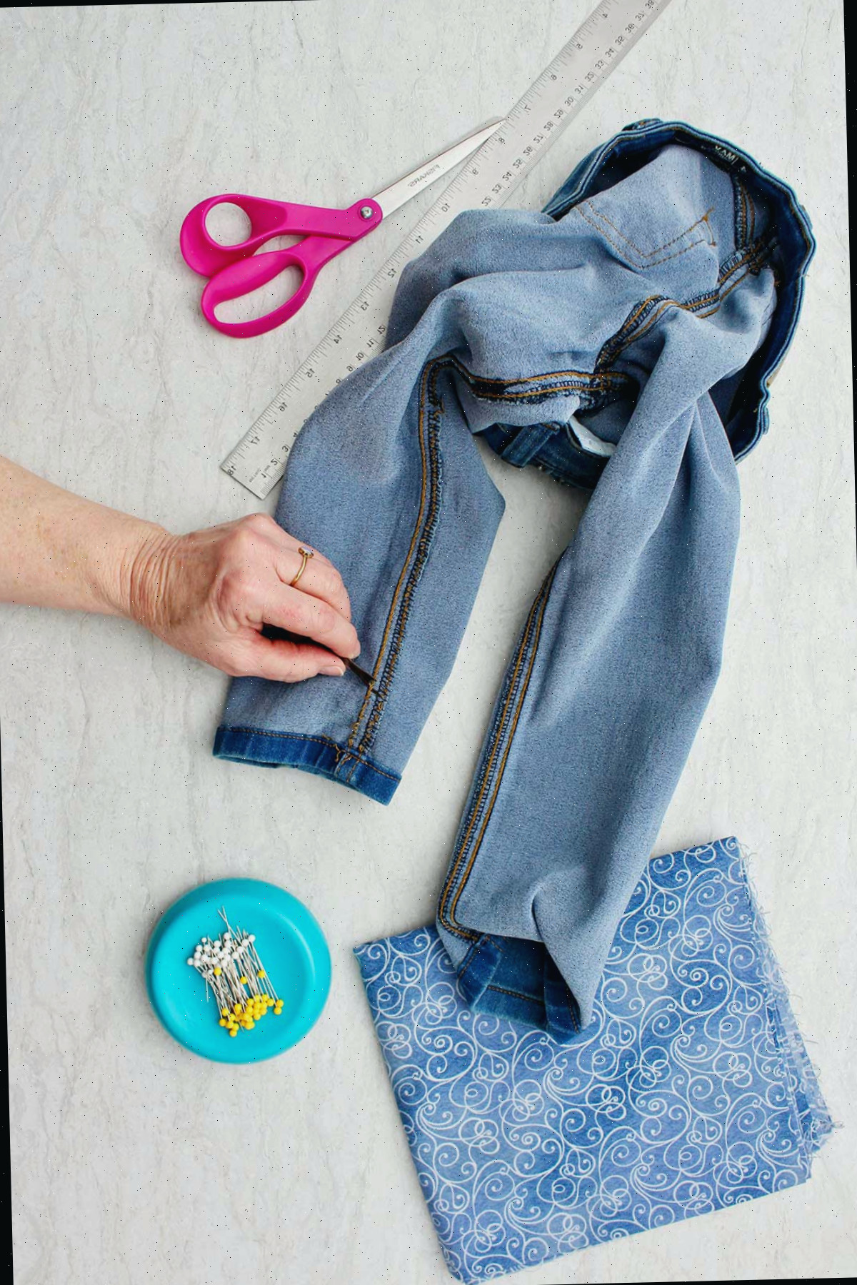 Um par de jeans do avesso, com a mão indicando onde deseja cortar para fazer barra sino, com alfinetes, tesouras e tecido ao redor.