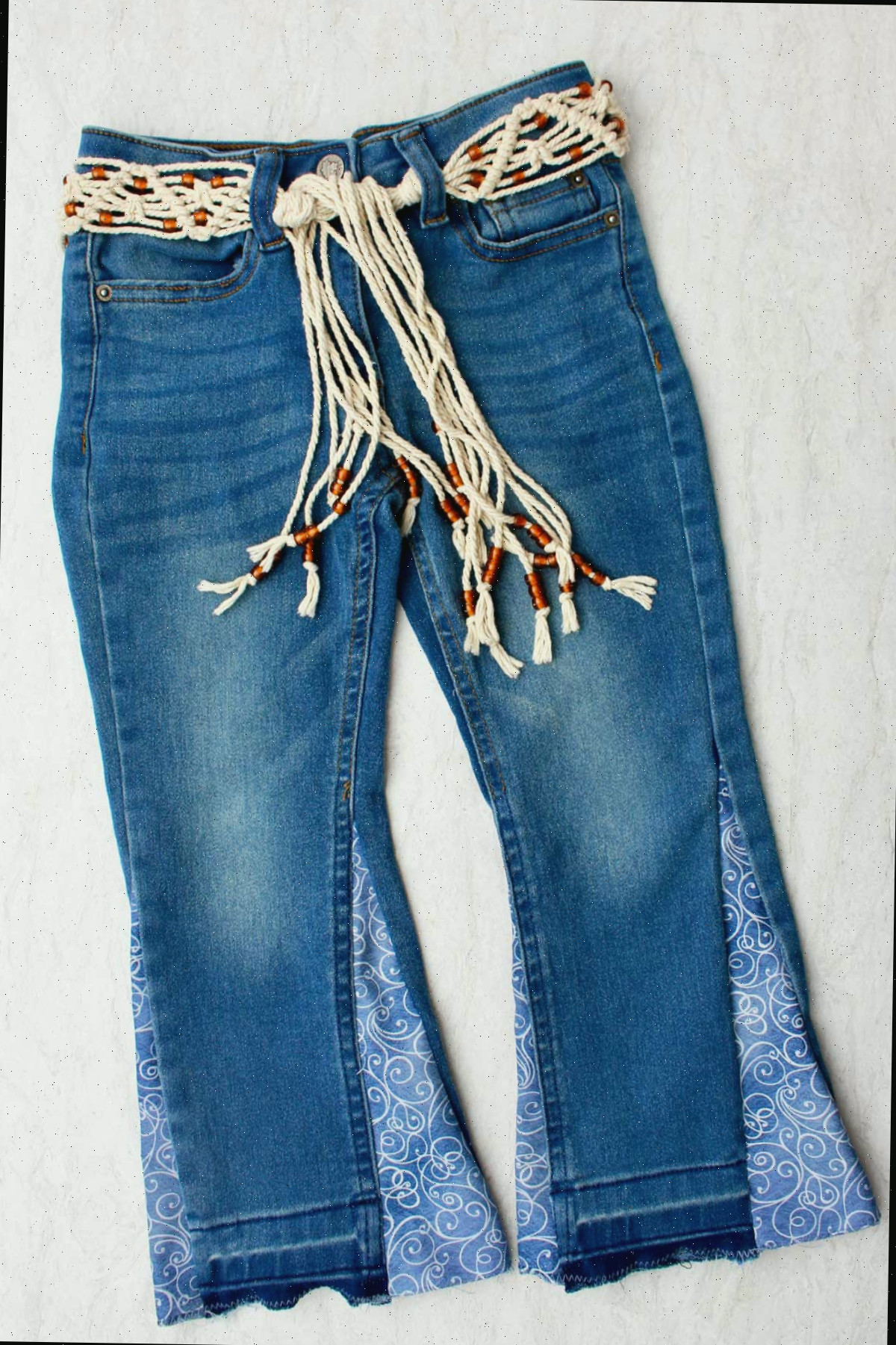 Um par de jeans infantis transformados em barra sino, com um tecido azul em espiral, preso com um cinto de macramê.
