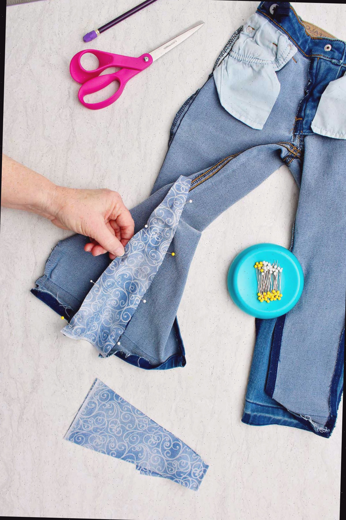 Mão segurando o tecido preso aos jeans com alfinetes retos, com suprimentos por perto.