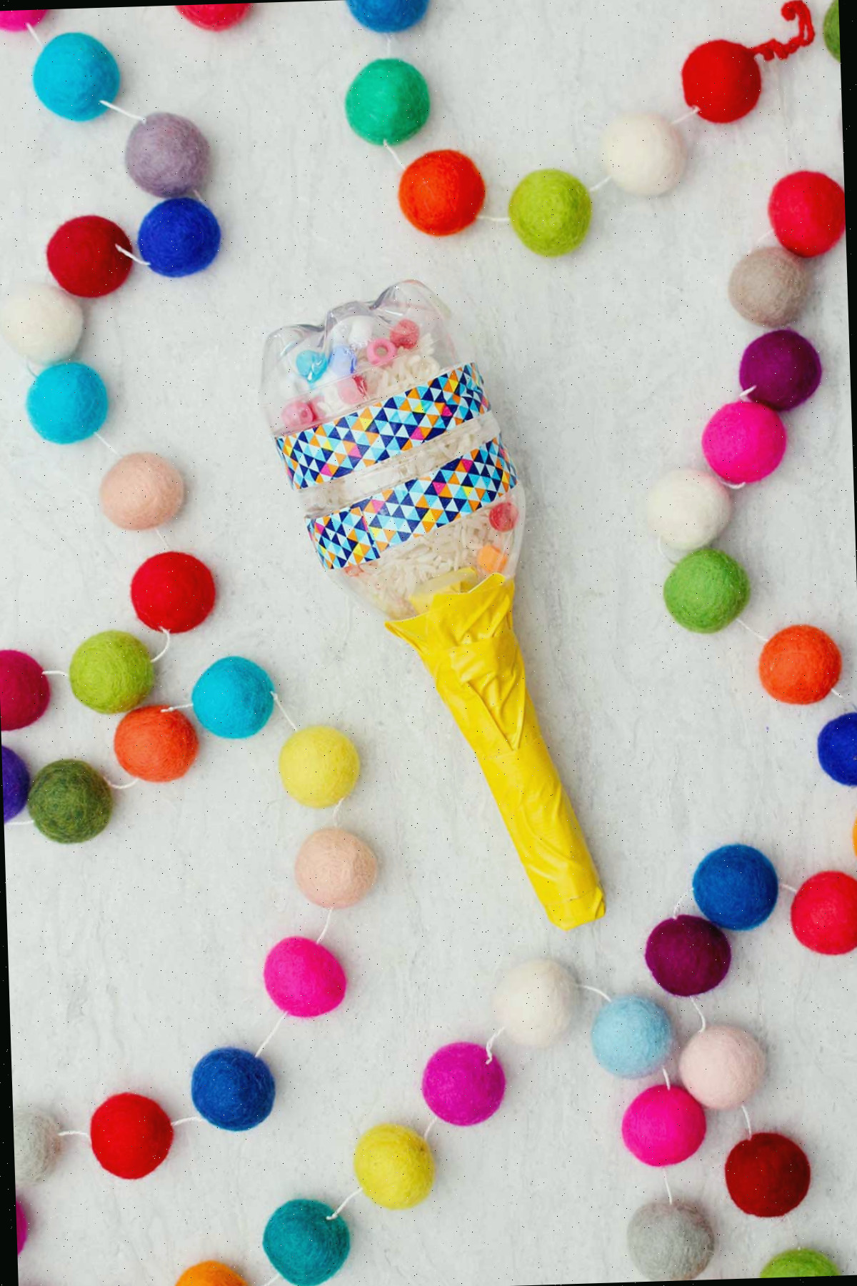 Maraca confeccionada com uma garrafa de água pequena preenchida com arroz e miçangas, com um cabo amarelo e pompons coloridos ao lado.