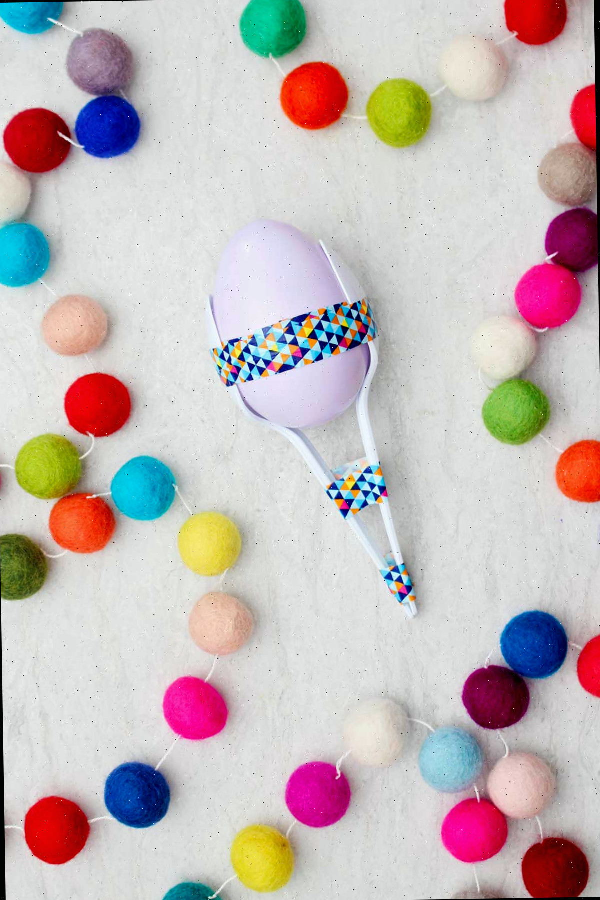 Maraca de ovo plástico lavanda com colheres e fita washi com pompons multicoloridos próximos.